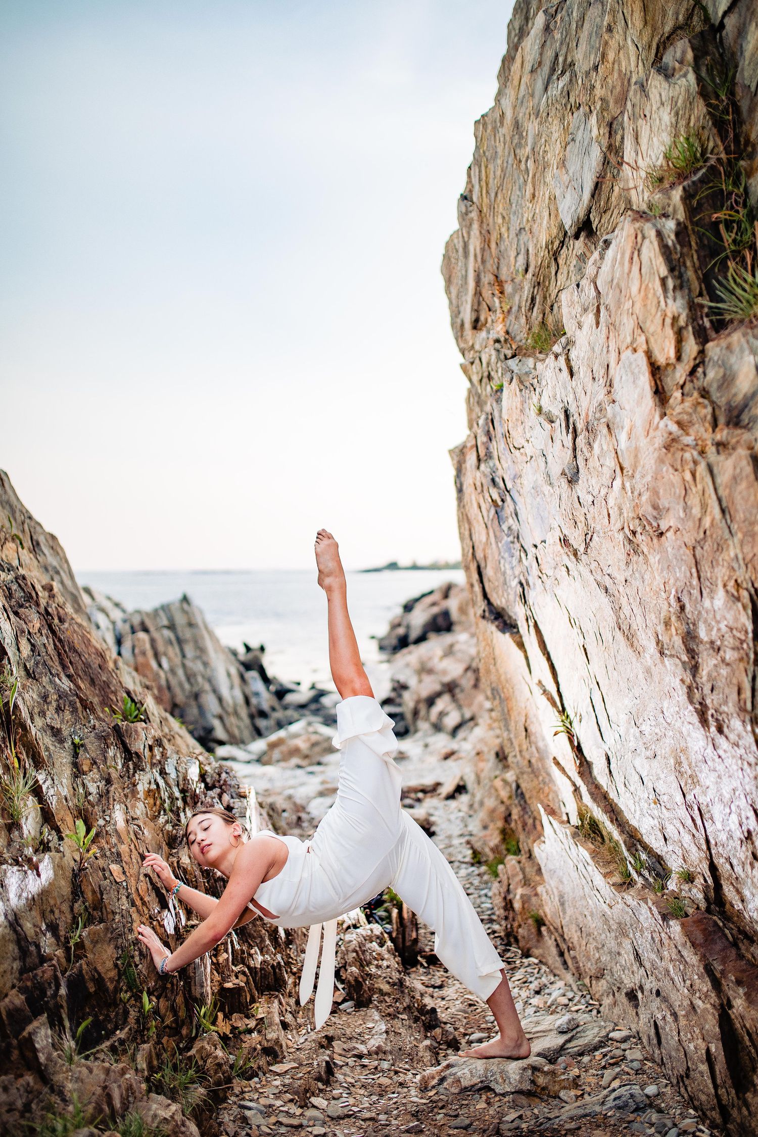 Woman in white outfit performing a dramatic acrobatic pose against a rocky cliff face near the ocean.