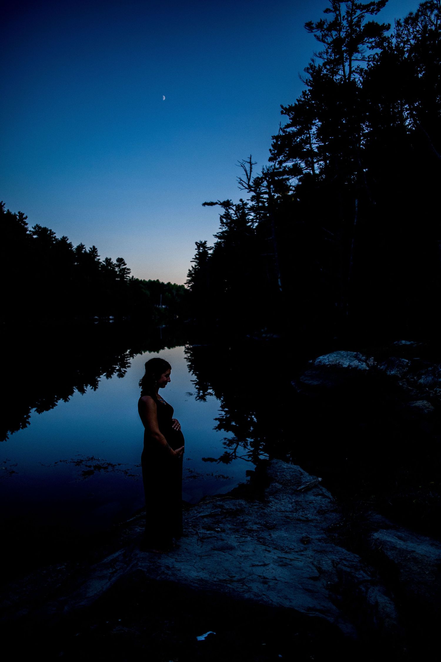 Silhouette of pregnant woman standing on rocks by reflective ocean cove at twilight under crescent moon.