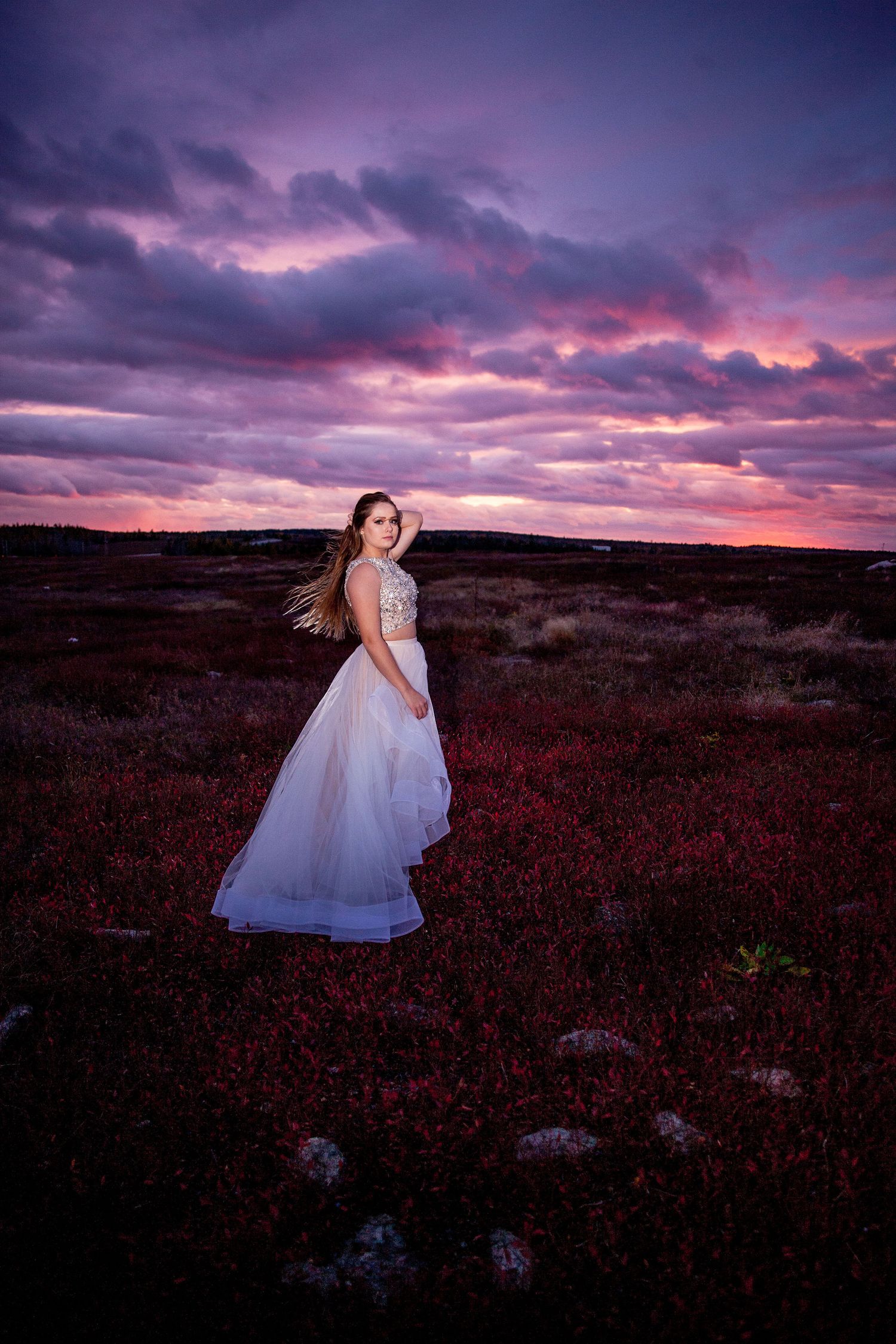 Young woman in white embellished gown stands in a field of red shrubs beneath a dramatic purple sunset sky.