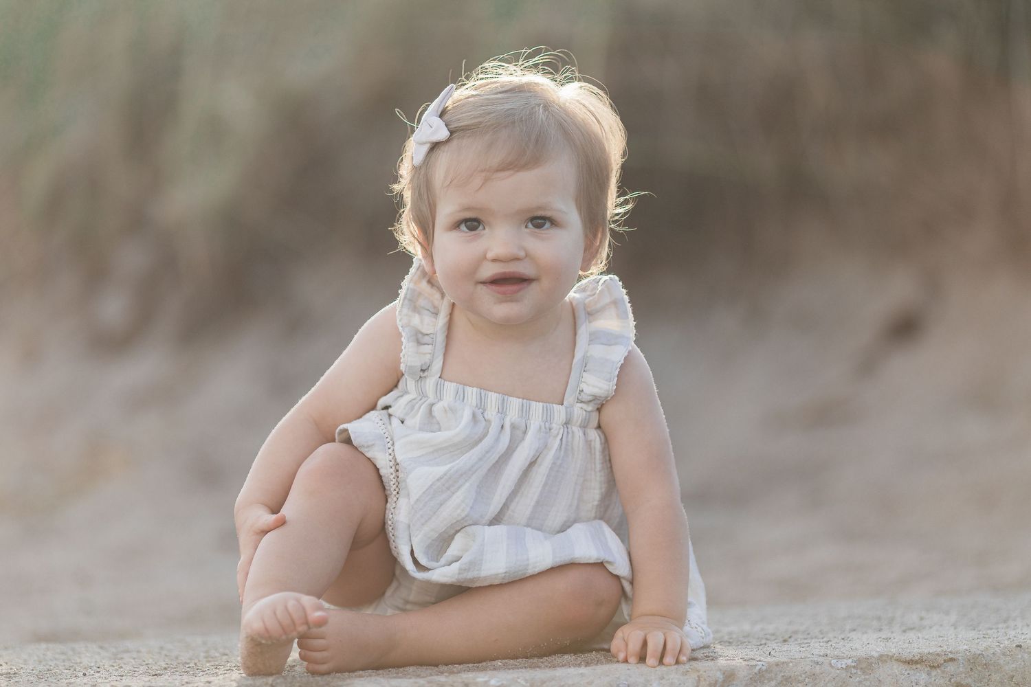 Cute baby girl in white ruffle romper sitting on sandy beach smiling in golden light.