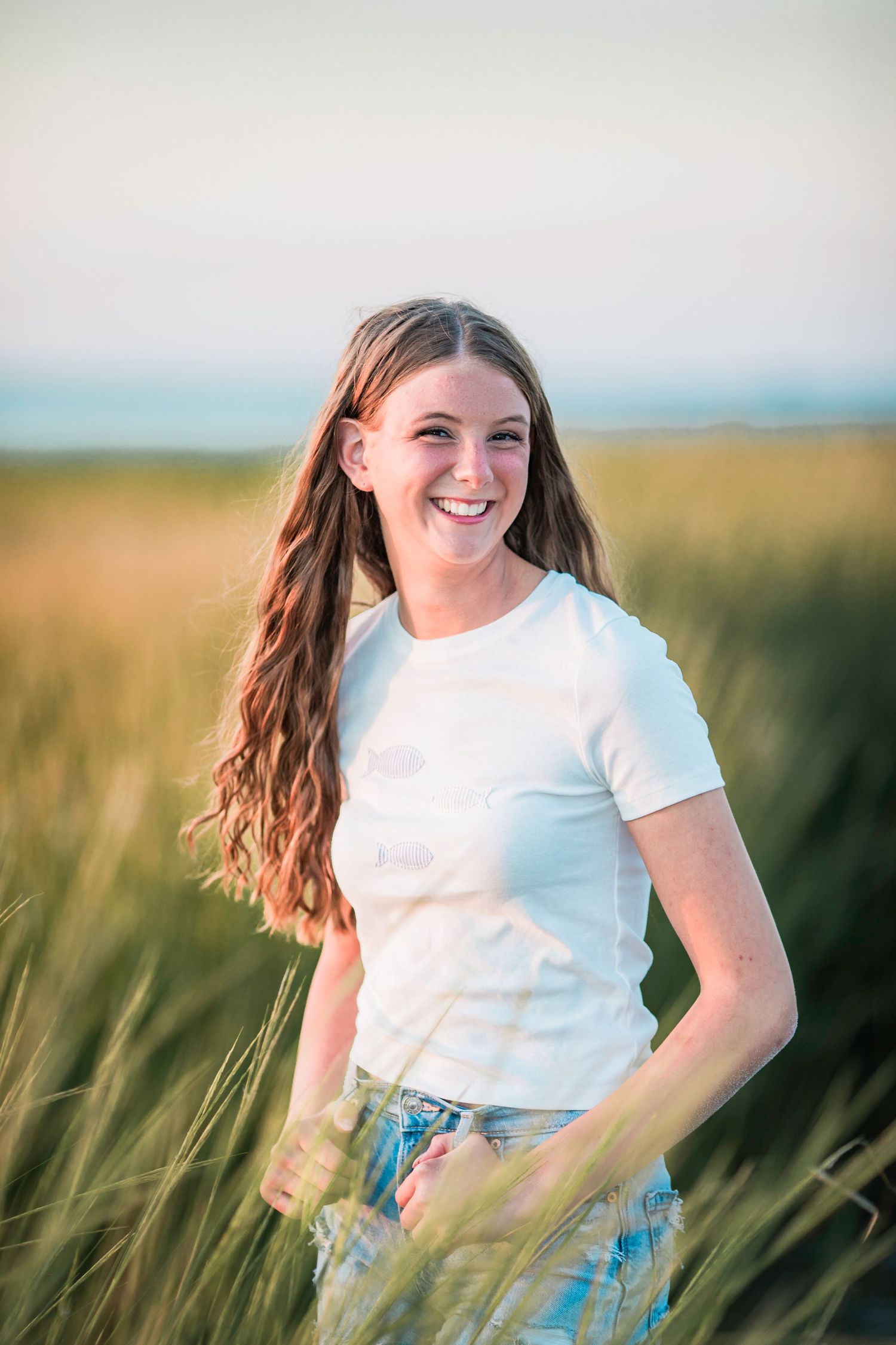 Smiling young woman with long auburn hair in white t-shirt and jeans standing in tall golden grass field.