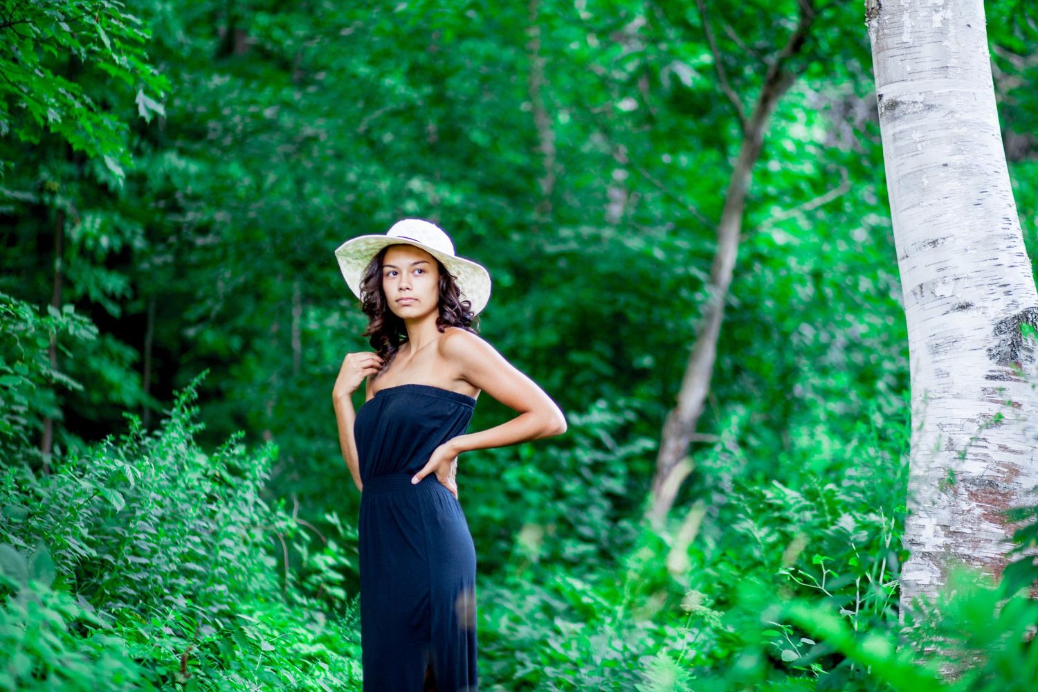 Young woman in black strapless maxi dress and white sun hat stands near birch tree in forest.