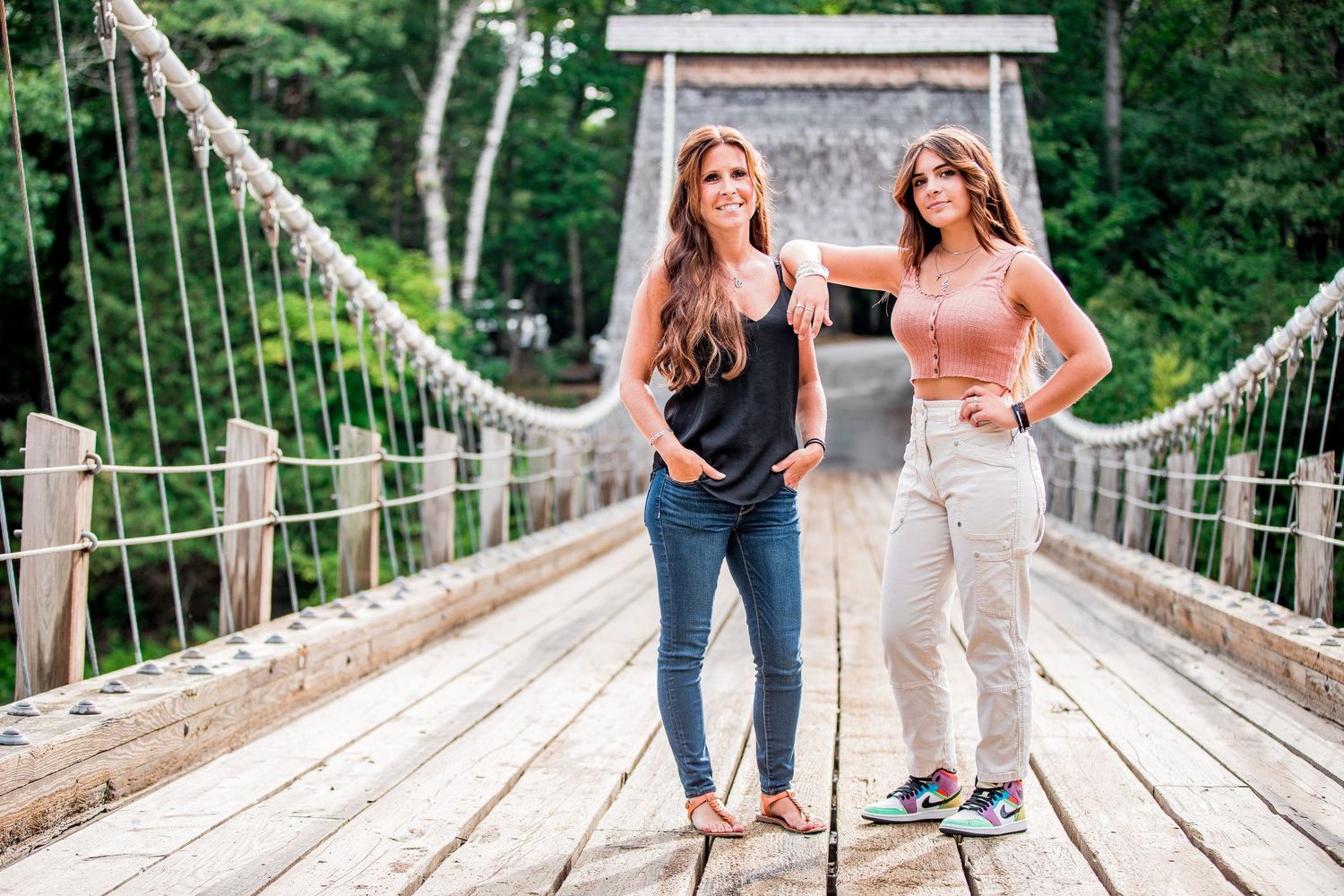 Two young women posing together on a wooden suspension bridge surrounded by trees.