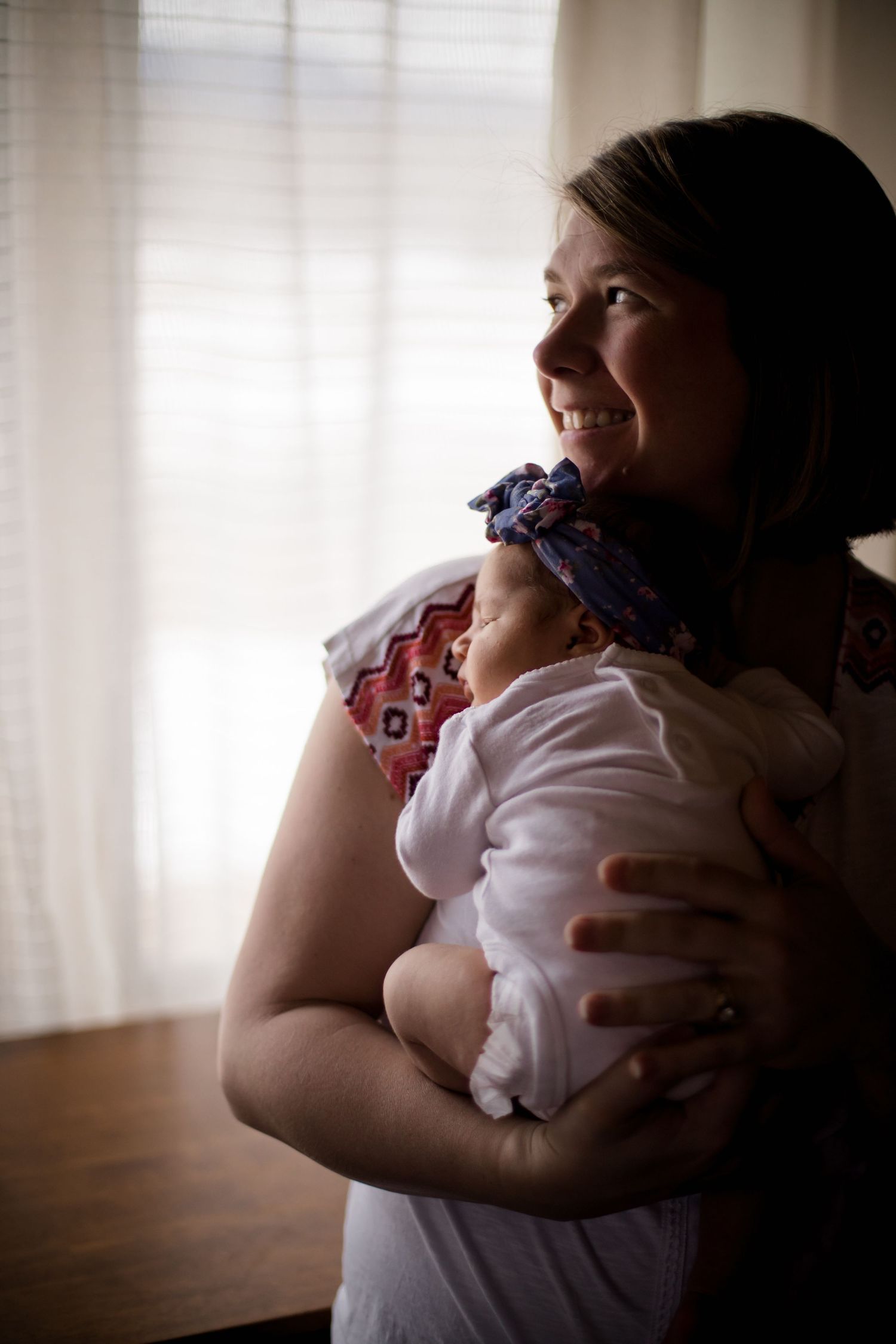 Happy smiling mother holding sleeping newborn baby with floral headband near sunlit window.