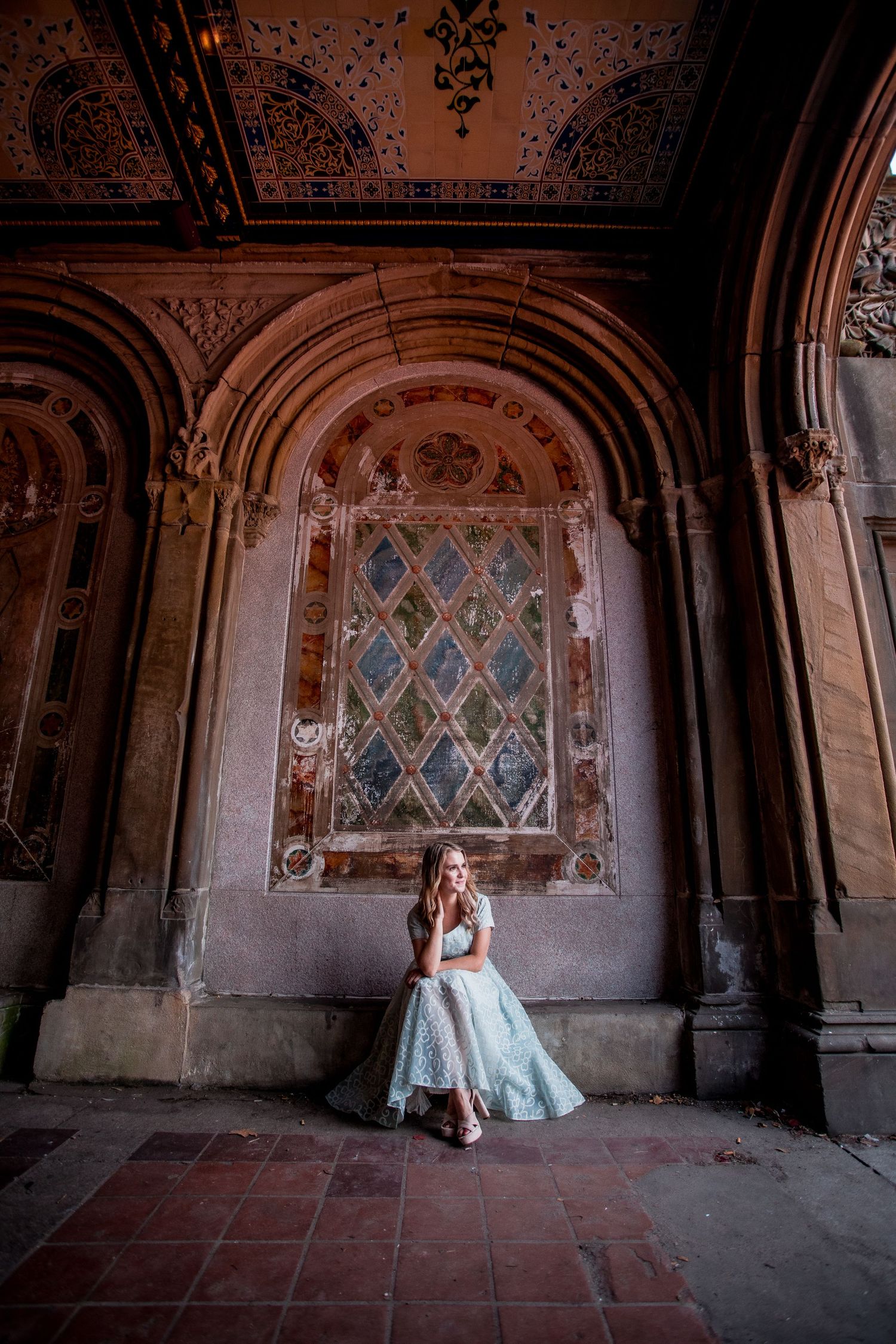 Young woman in light blue tulle dress sitting inside ornate Bethesda Terrace arcade in Central Park.