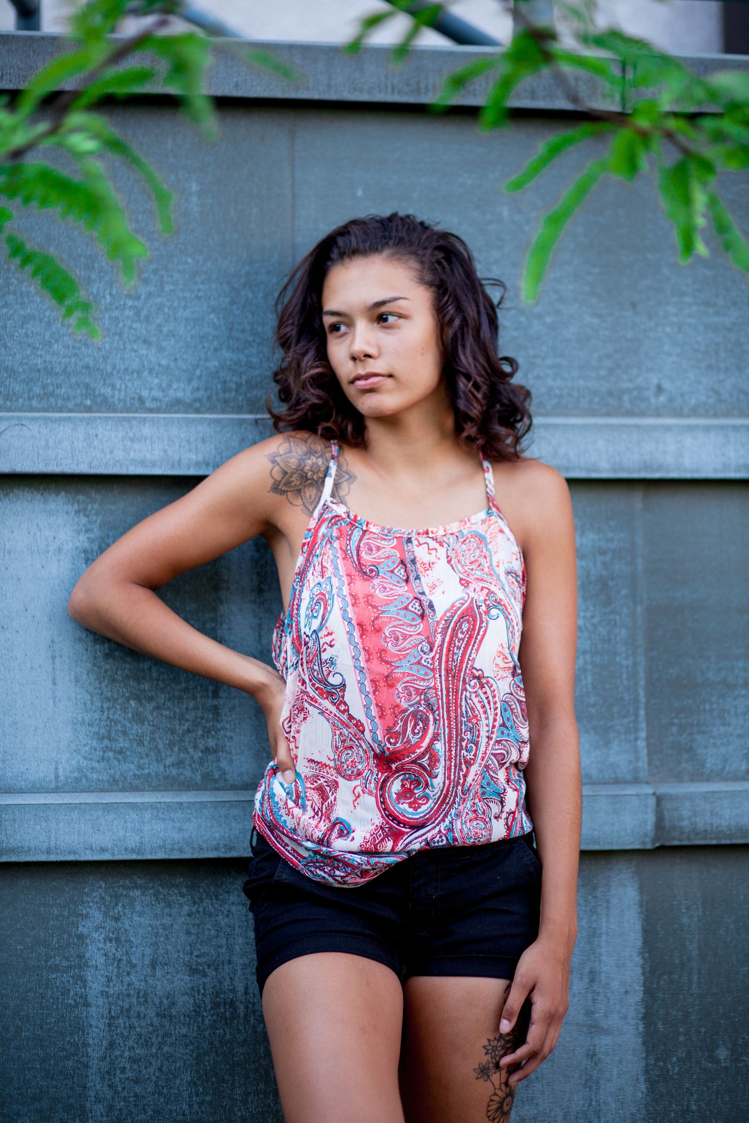 Young woman in paisley tank top and black shorts leaning against a metal wall outdoors.