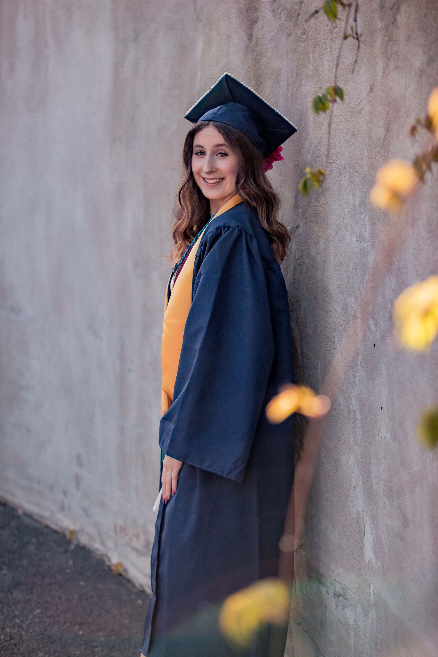 Smiling graduate in navy cap and gown with gold stole posing against a concrete wall outdoors.