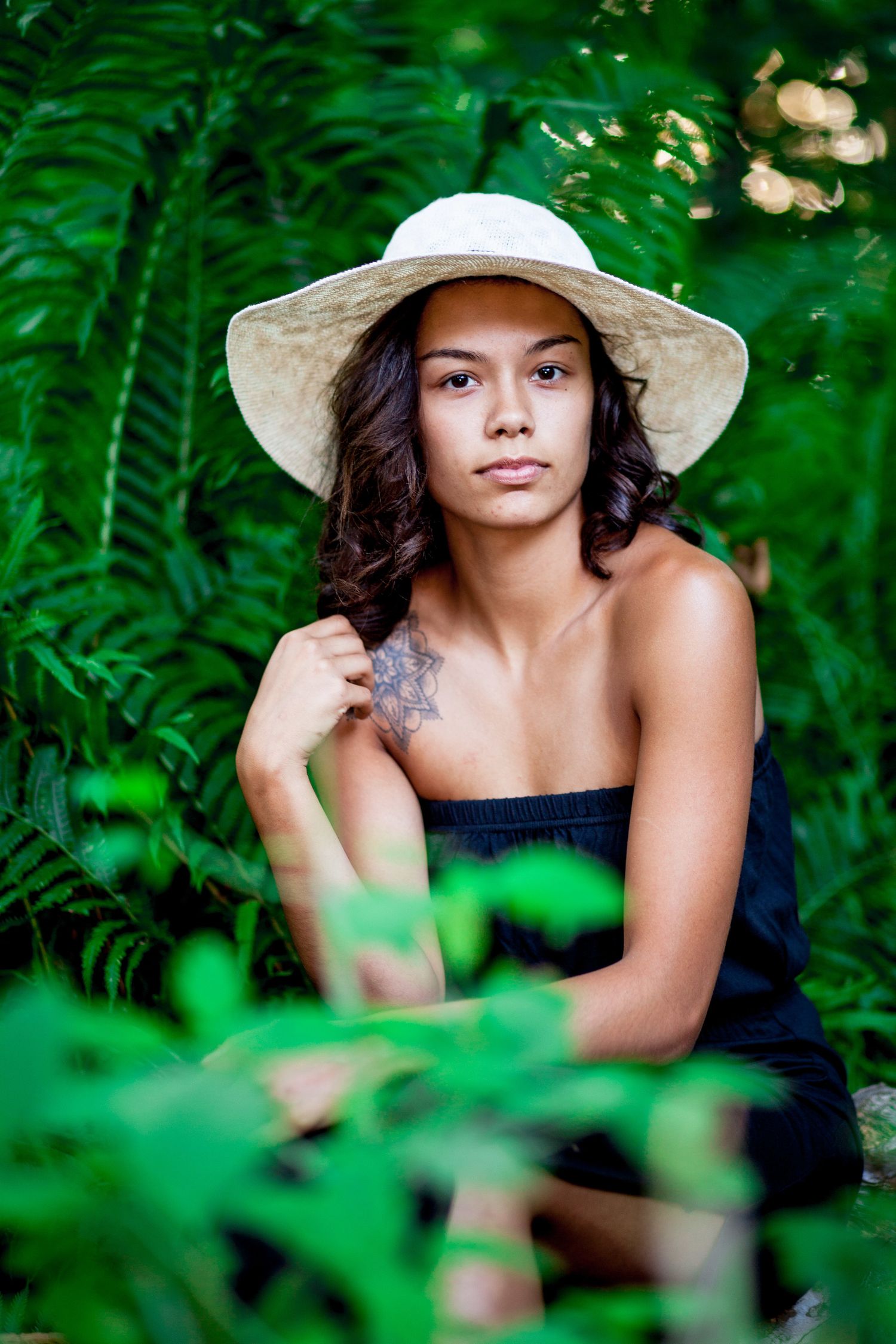 Young woman with tattoo wearing white sun hat poses among lush green tropical ferns outdoors.