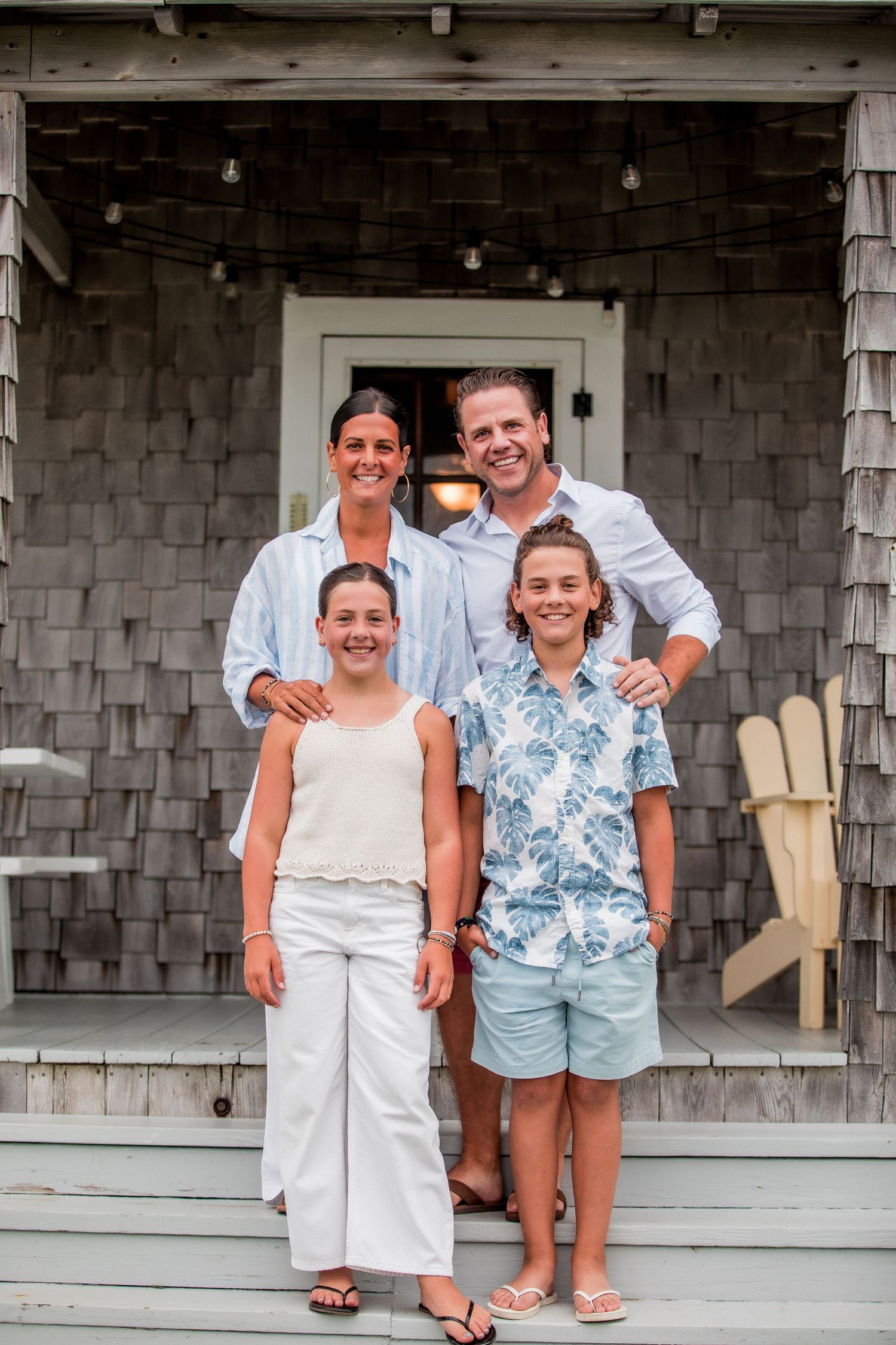 Family of four posing on porch steps of shingled beach house in summer outfits.