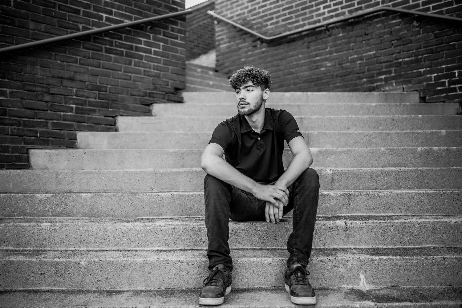 Black and white photo of a young man sitting on outdoor concrete steps near brick wall.