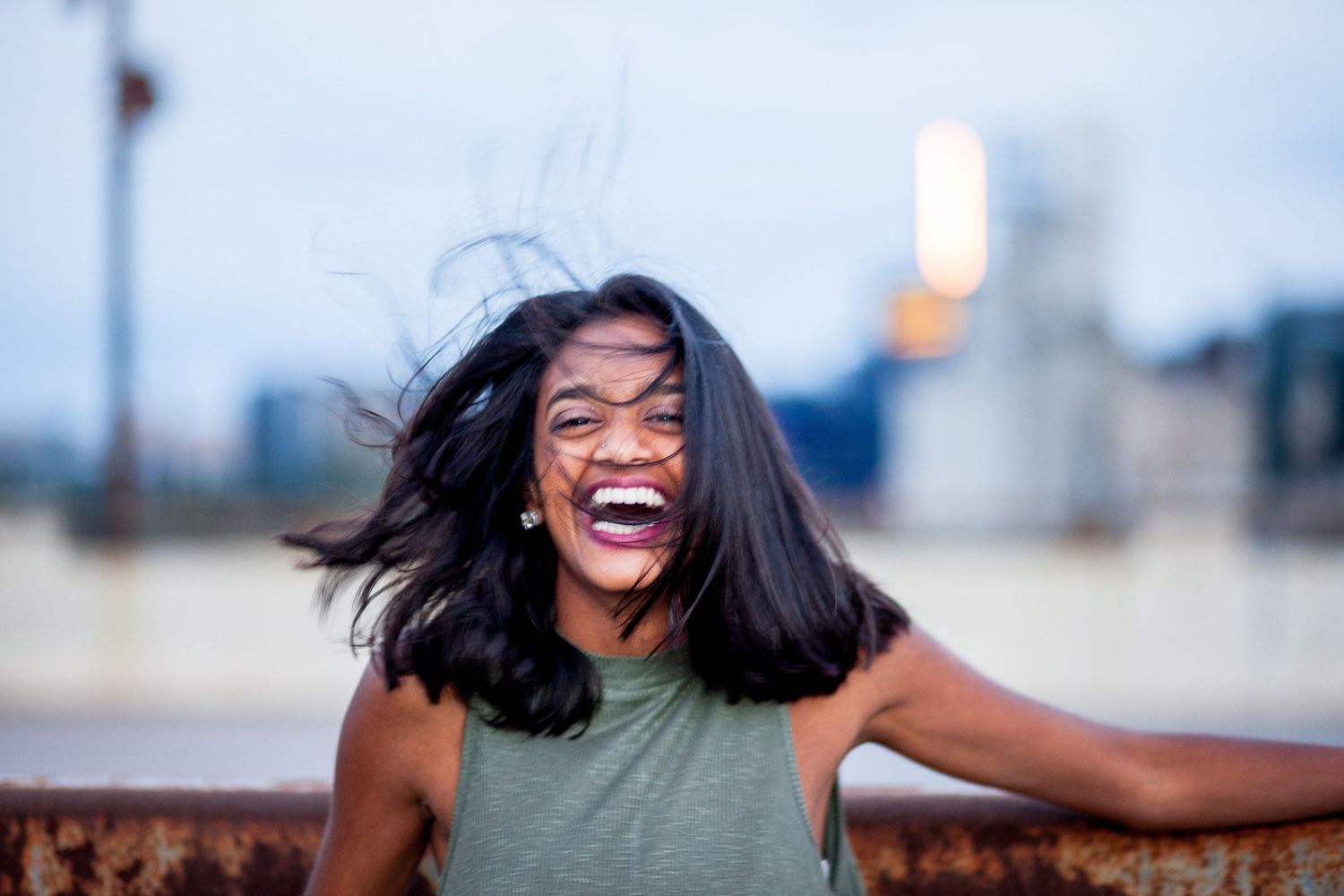 Joyful Black woman in green sleeveless top laughs with hair blowing in wind against city backdrop.
