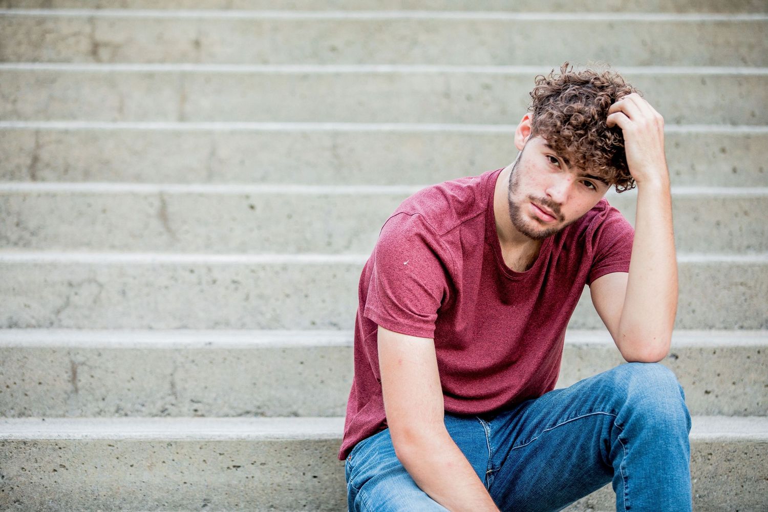Young man with curly hair in a red t-shirt sitting on concrete steps outdoors.