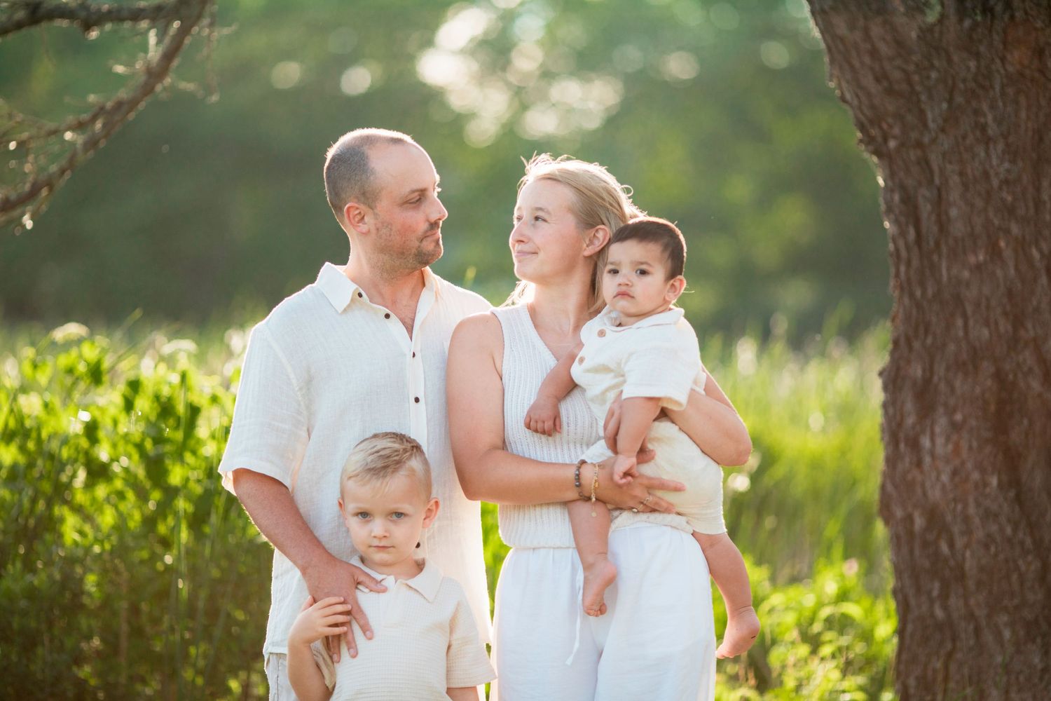 Family of four in white outfits posing outdoors among green trees in warm golden sunlight.