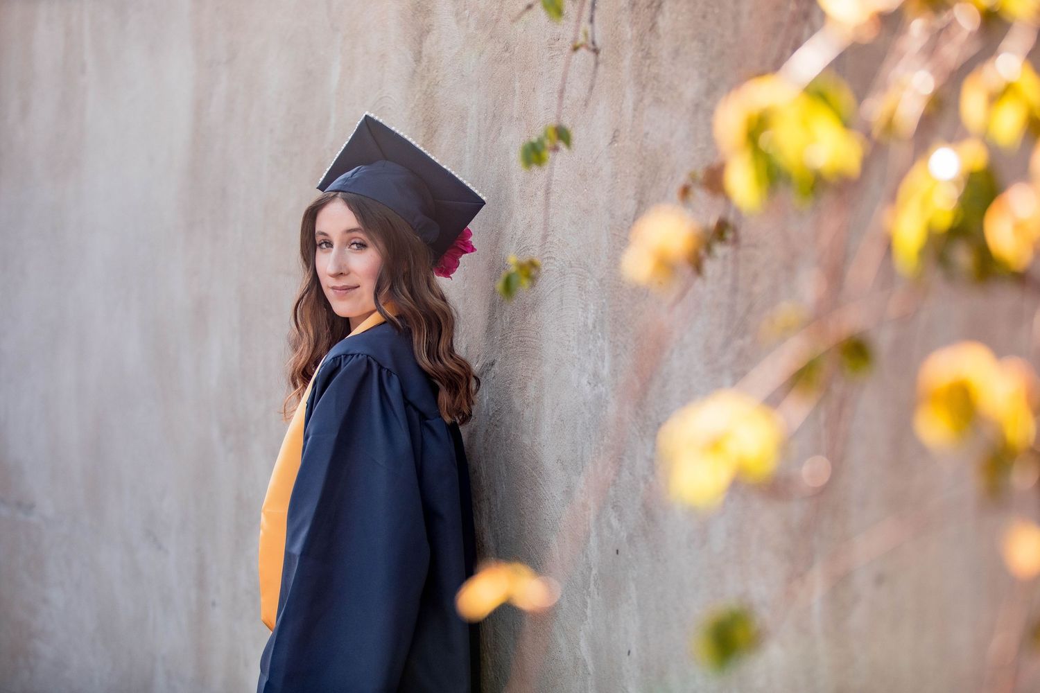 Young woman in graduation cap and gown posing against a concrete wall with autumn foliage in background.