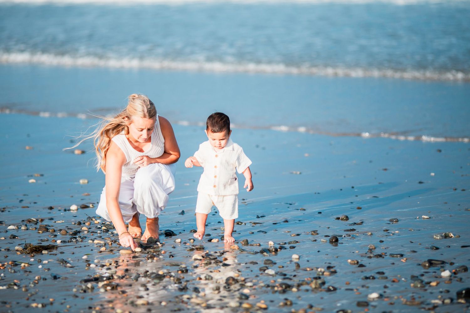 Woman and toddler in white outfits playing on a sandy beach near ocean waves on a sunny day.
