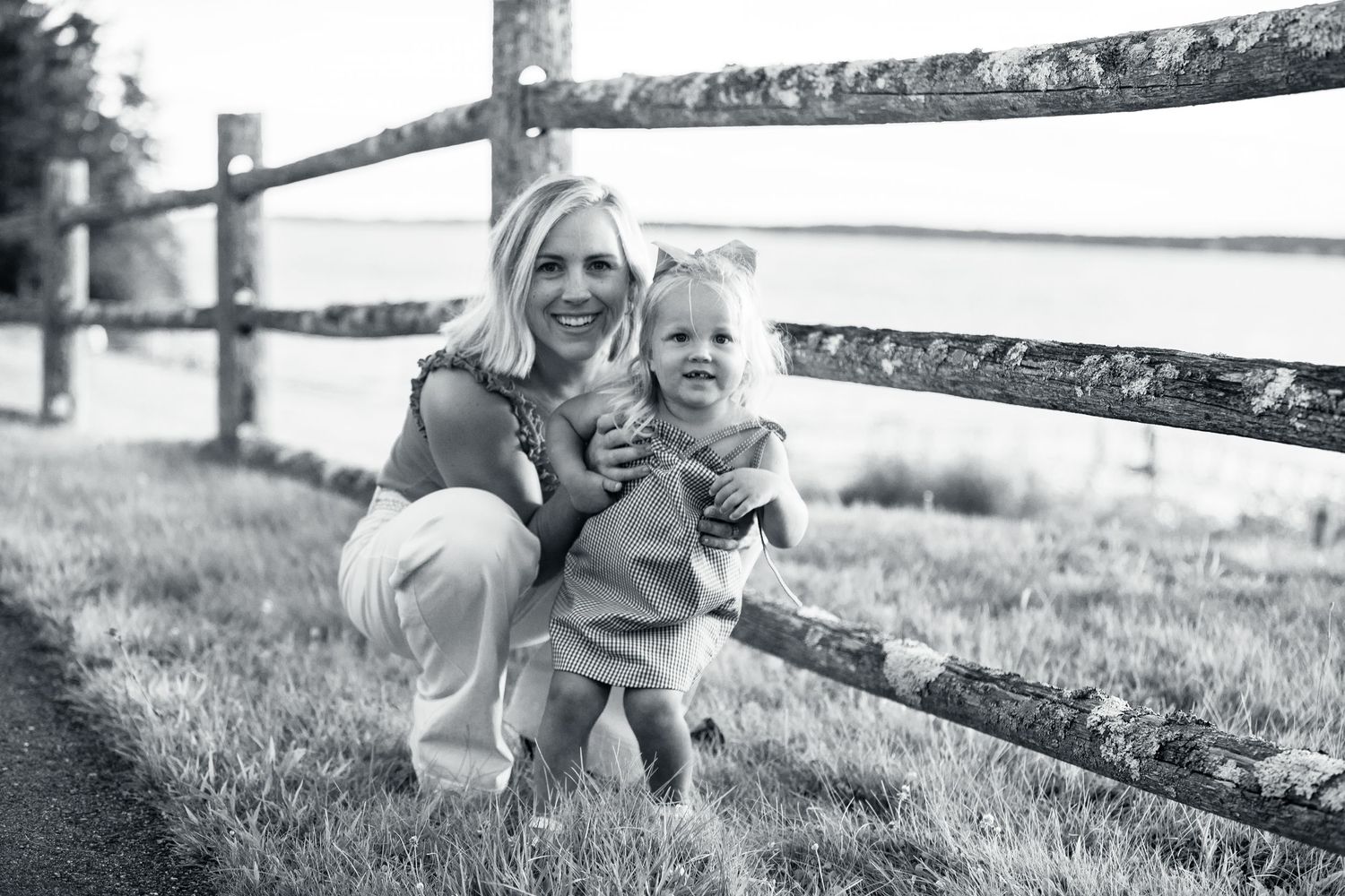 Black and white photo of smiling mother crouching with toddler by wooden fence outdoors by the Cape Cod ocean.
