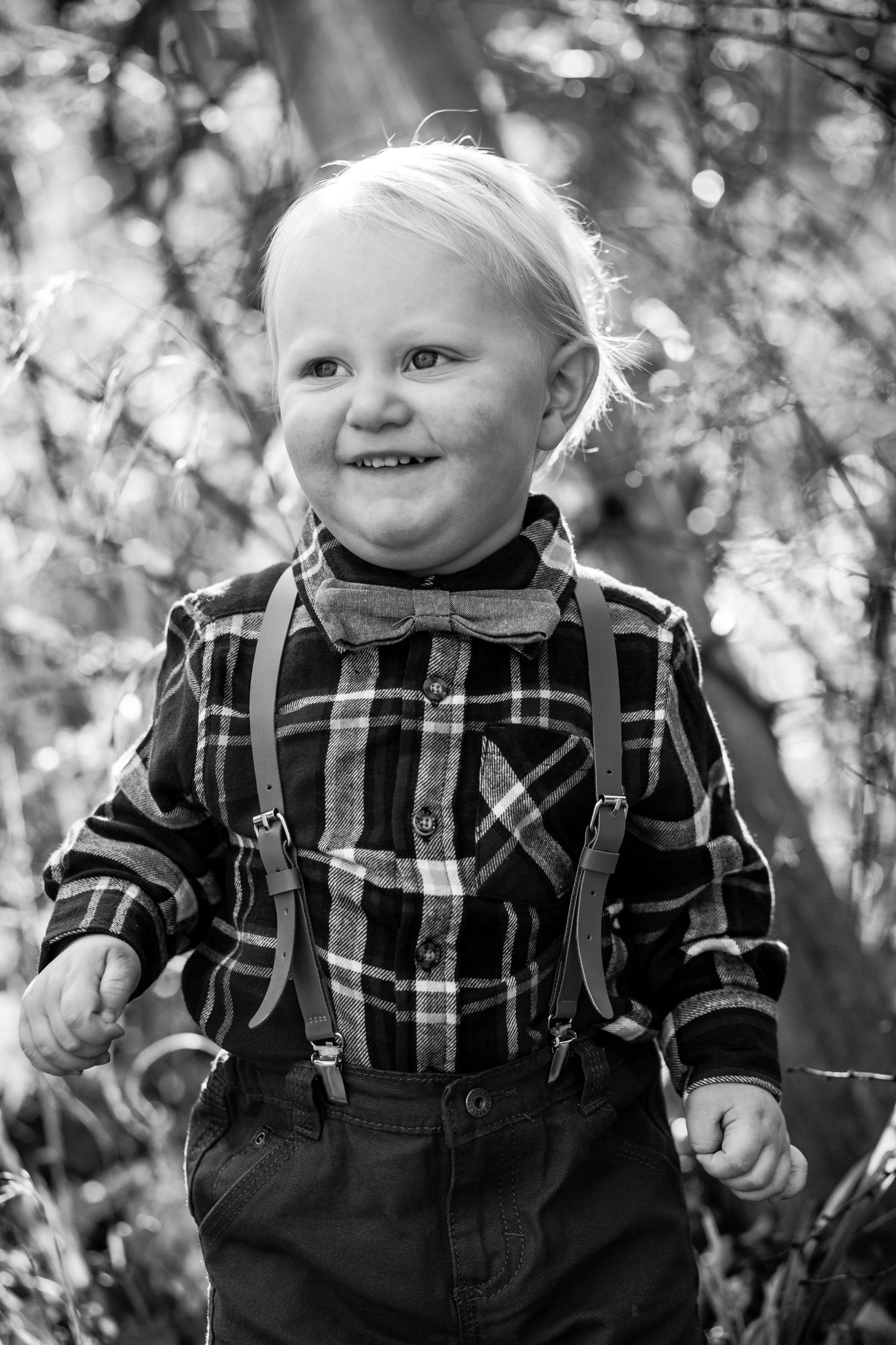 Happy toddler boy in plaid shirt with bow tie and suspenders smiling in black and white outdoor photo.