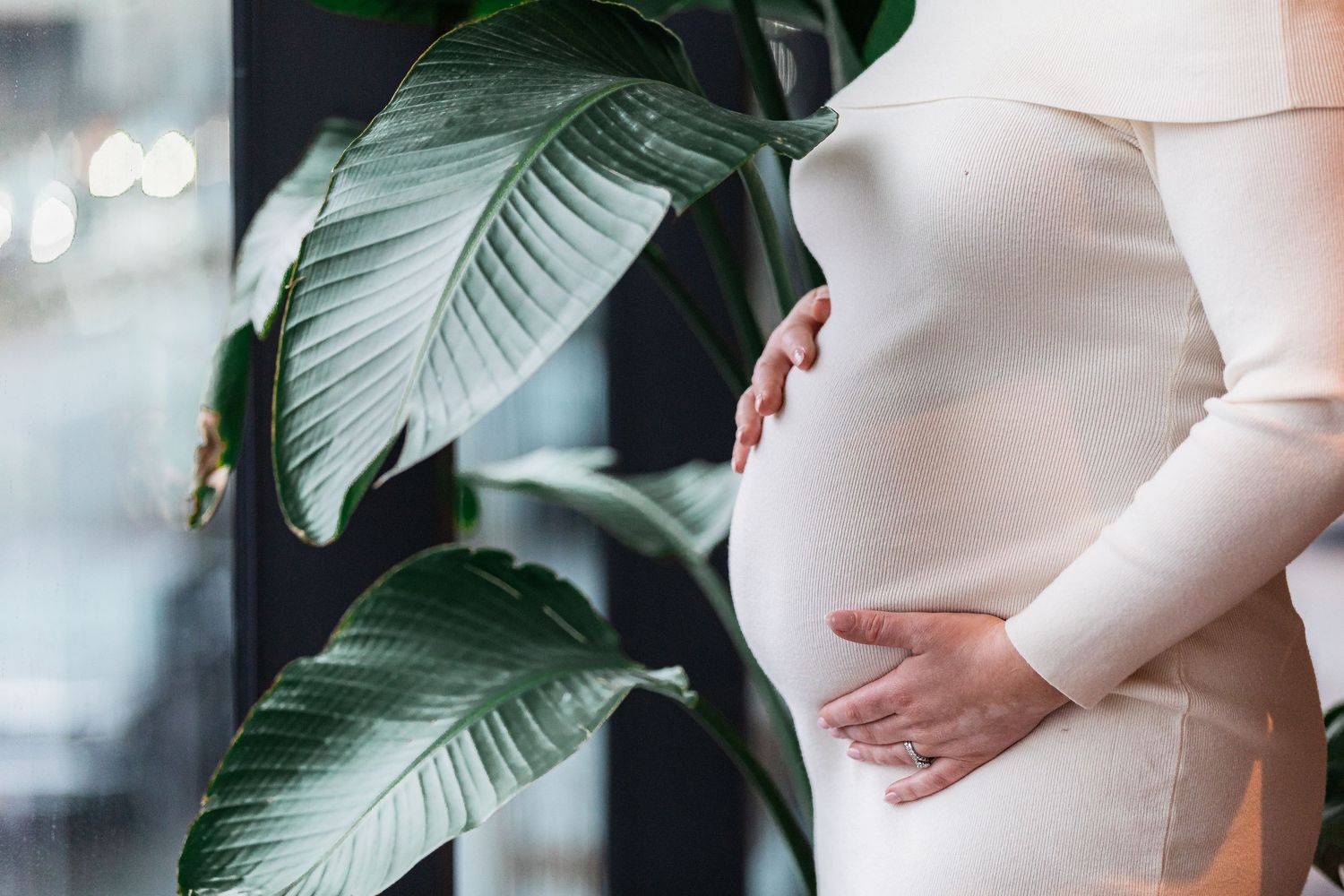 Close-up of pregnant woman in white dress cradling baby bump near tropical plant.