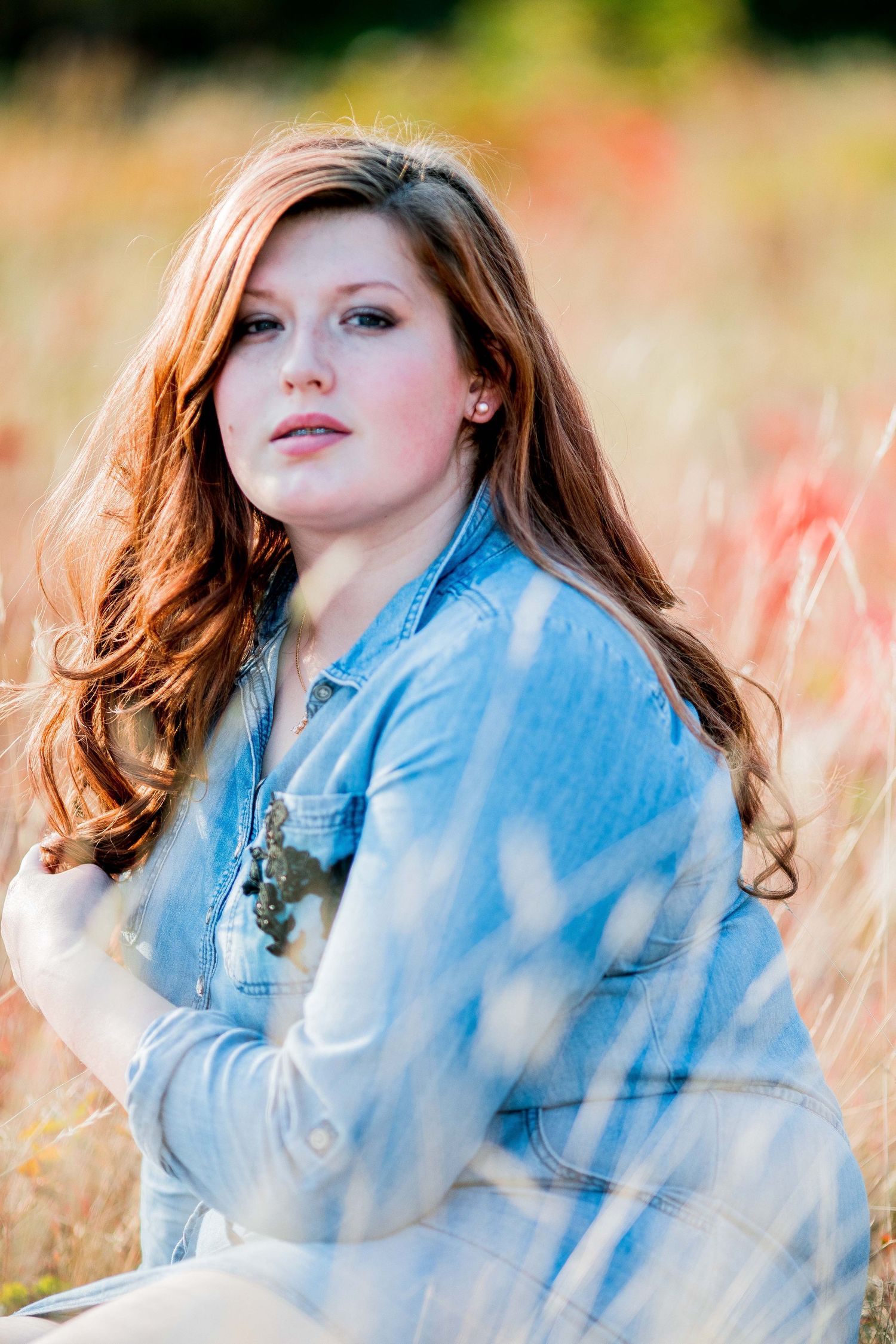Young woman with red hair sitting in a golden meadow wearing a blue denim shirt.