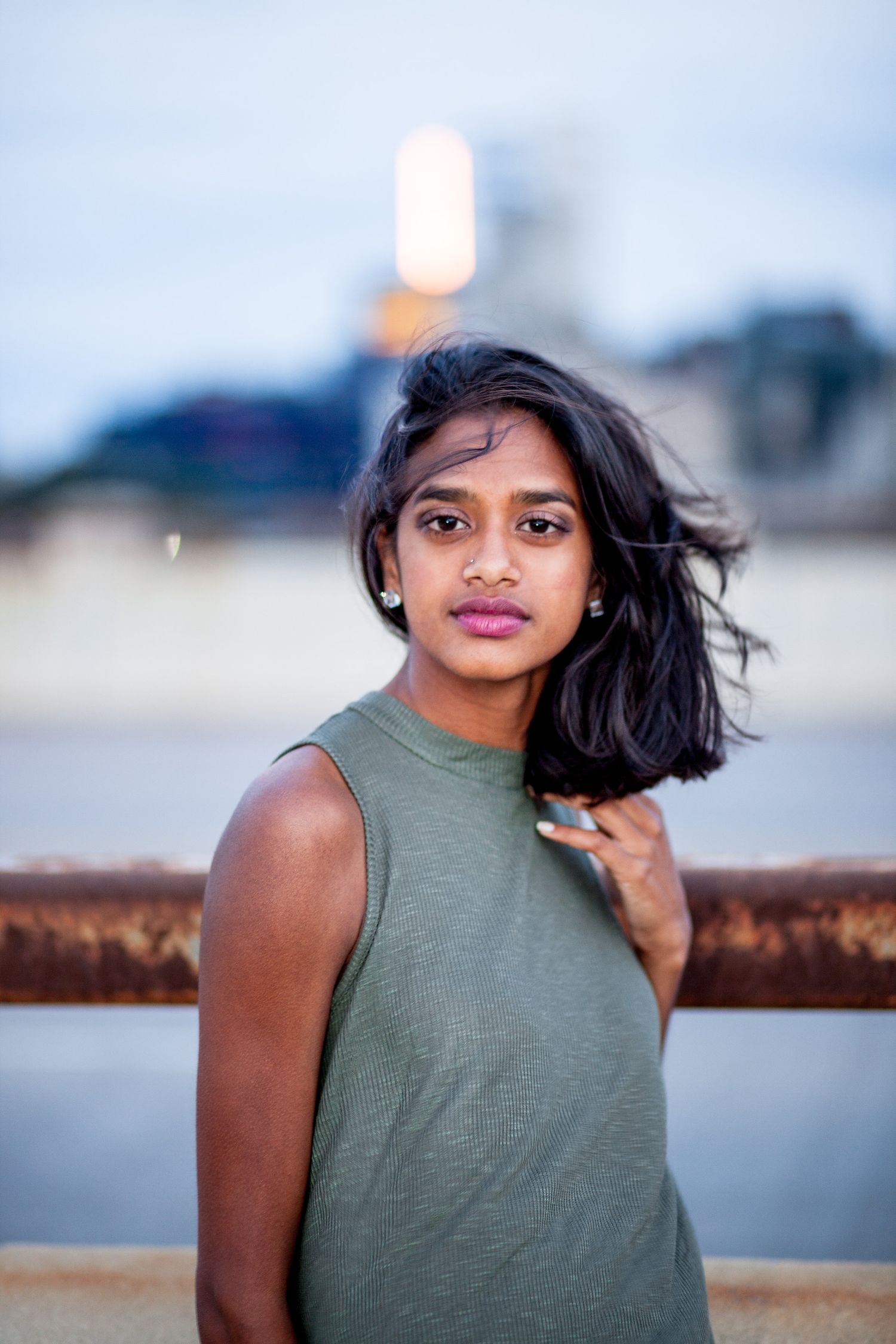 Young South Asian woman in olive sleeveless top poses outdoors near rusty railing with wind-blown hair.
