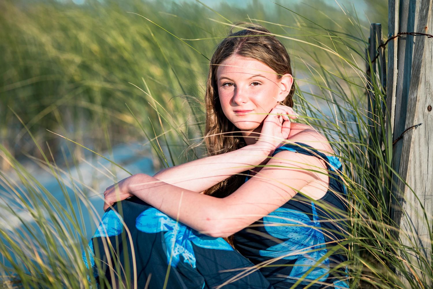 Young girl in blue dress poses among tall beach grass near a wooden fence in a sunny outdoor portrait.