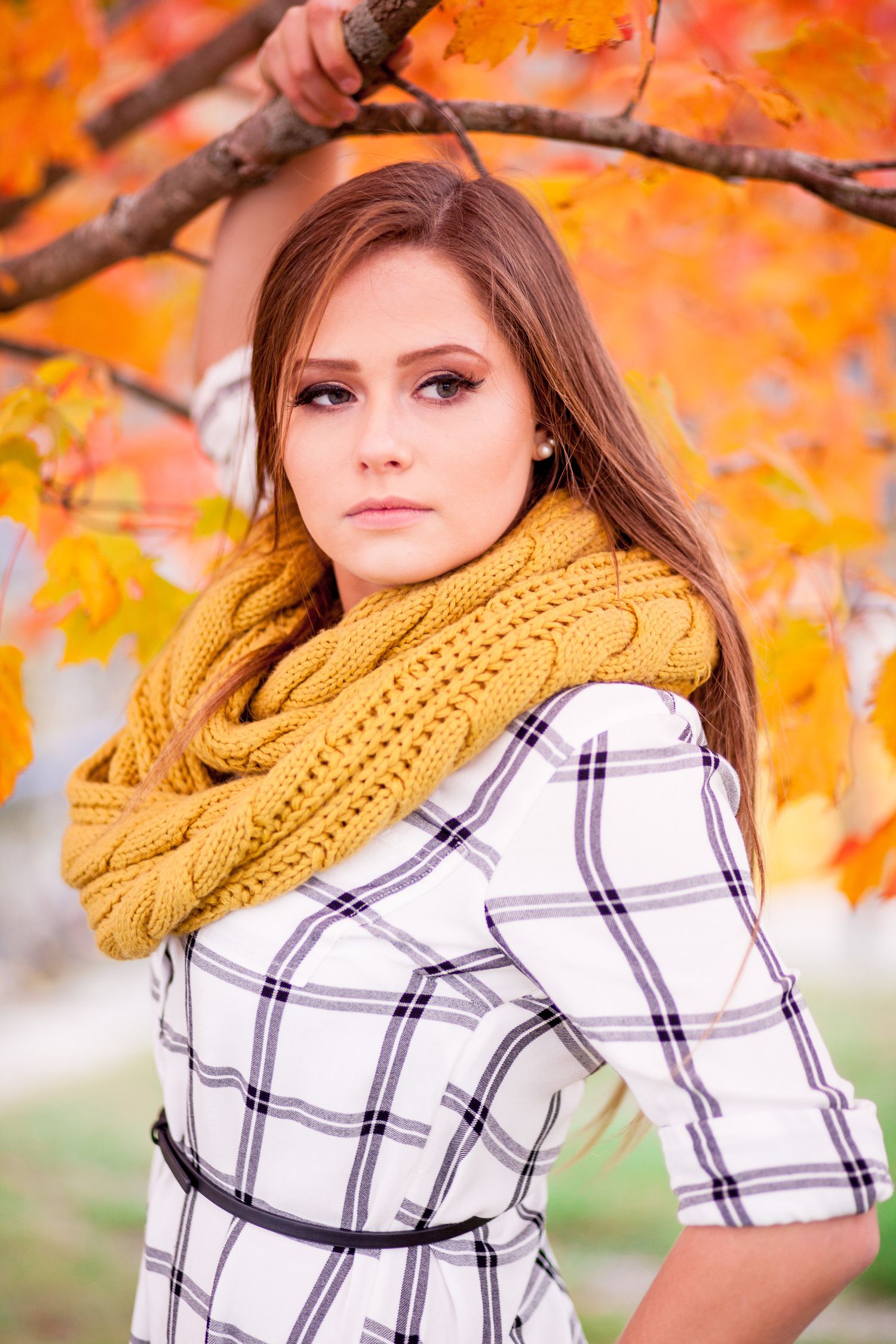 Close-up portrait of a young brunette woman in plaid dress and yellow knit scarf among fall foliage.
