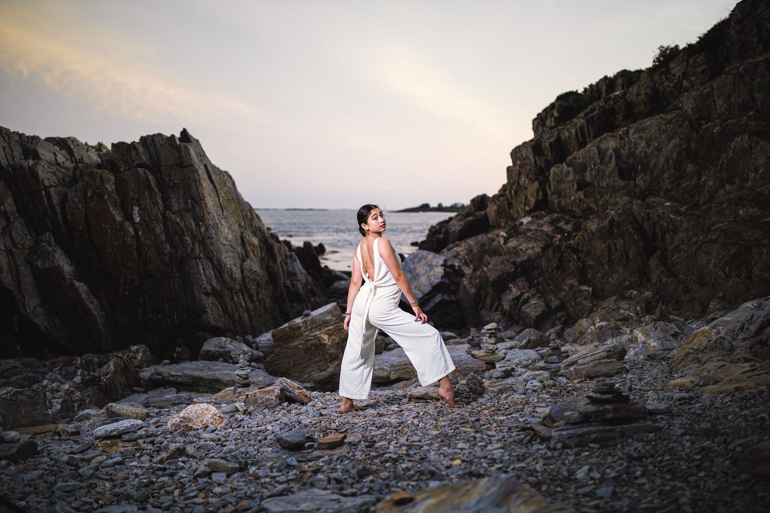 Woman in white wide-leg pants standing among large rocky boulders near the ocean at dusk.