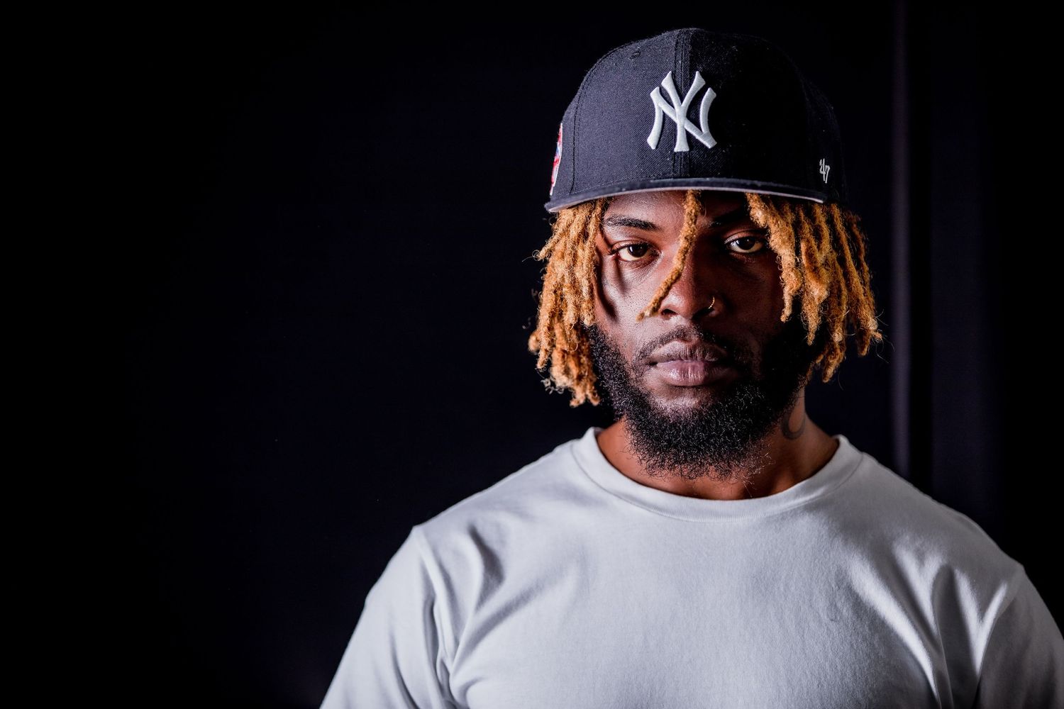 Confident man with dreadlocks wearing a New York Yankees cap and white t-shirt on dark background.