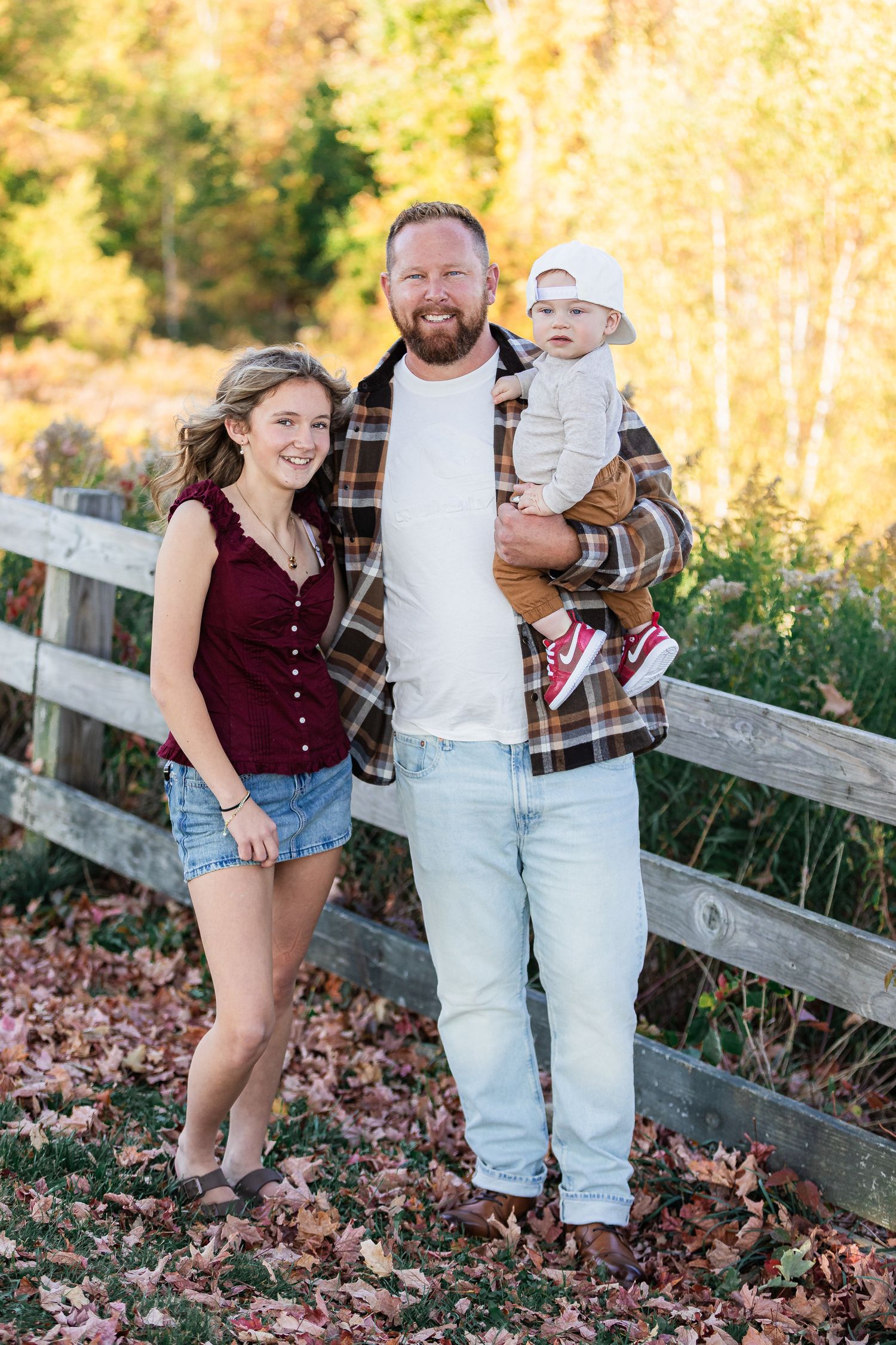 Smiling man holding baby with teenage girl beside wooden fence surrounded by colorful autumn foliage.