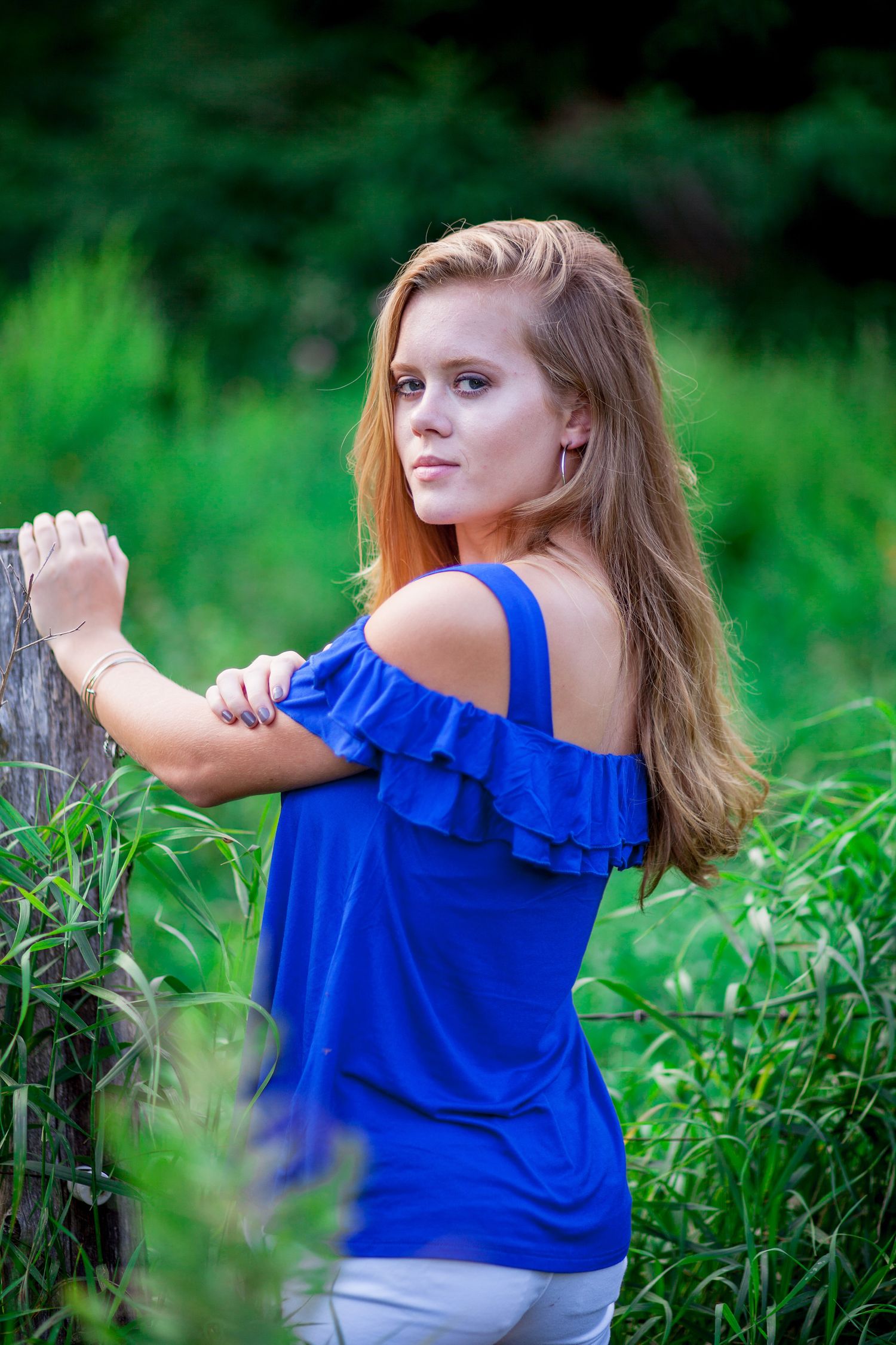 Young woman in blue off-shoulder ruffled top standing by a wooden post in lush green outdoor setting.