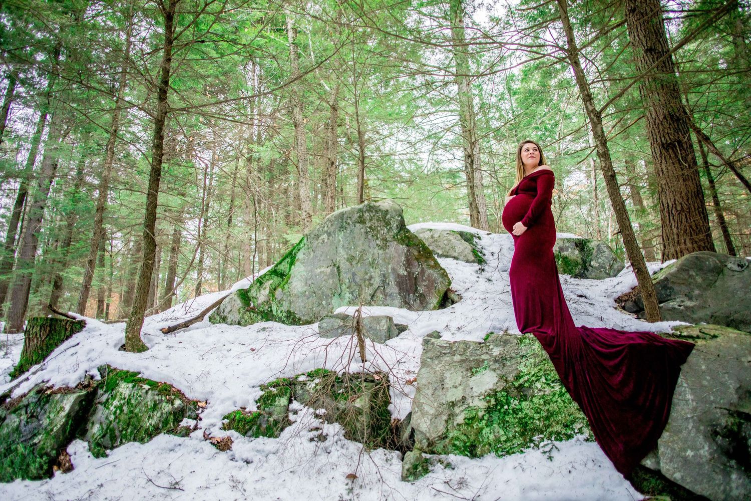Pregnant woman in red gown standing on snowy rocks in winter forest.