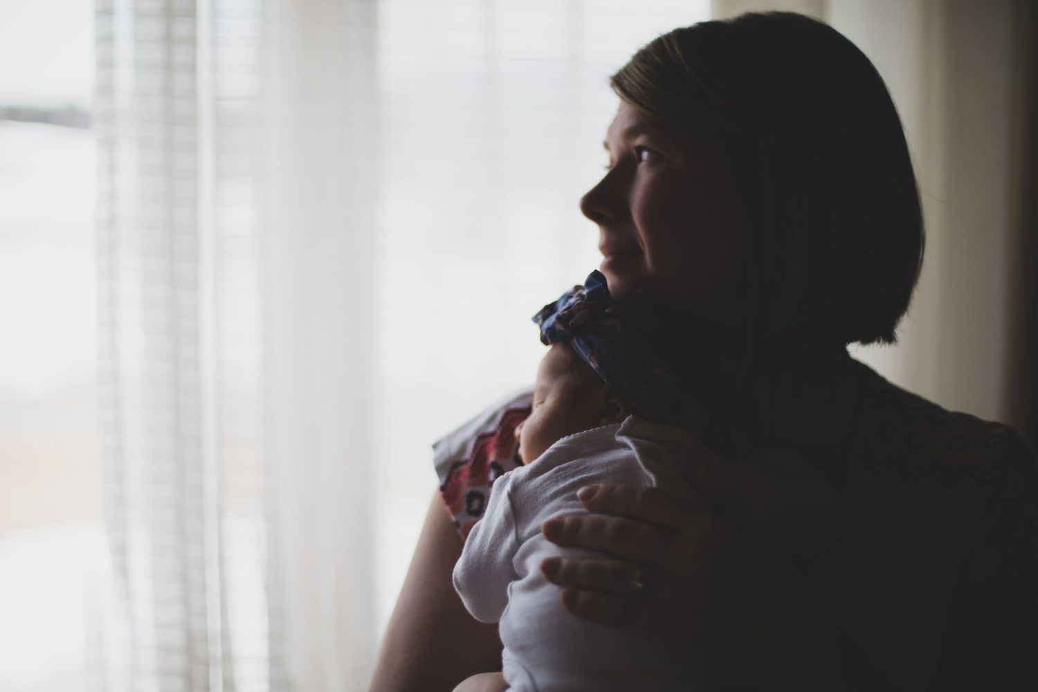 Silhouette of mother holding newborn baby near bright window with soft curtains in background.
