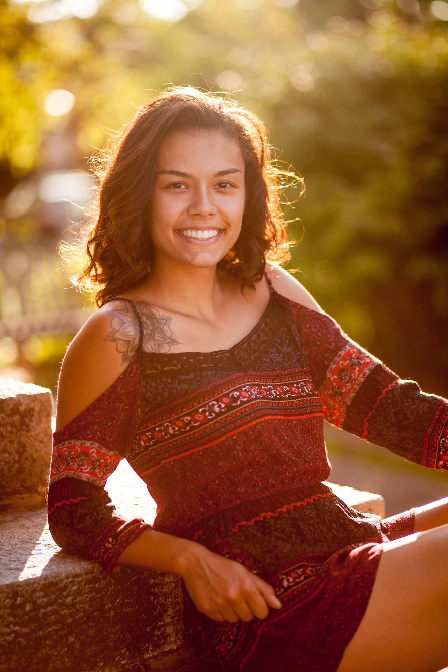 Smiling young woman in red boho off-shoulder top sitting outdoors in warm golden sunlight.