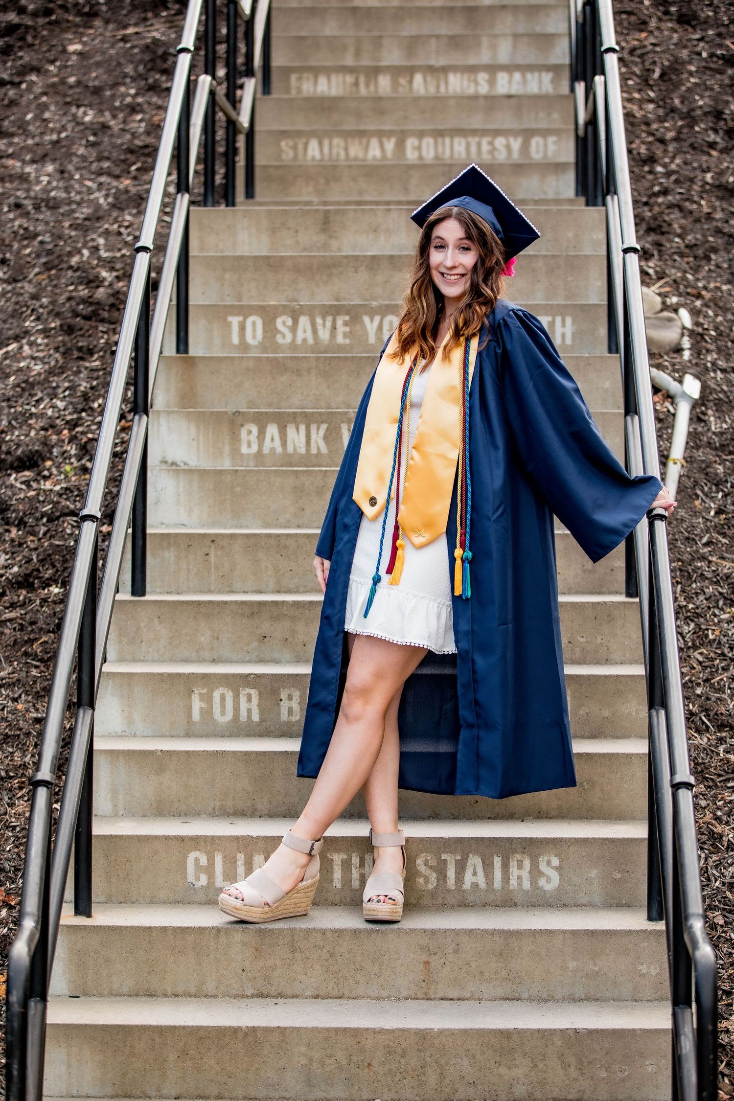 Graduate in navy cap and gown with gold stole posing on stone stairs with inscribed text.