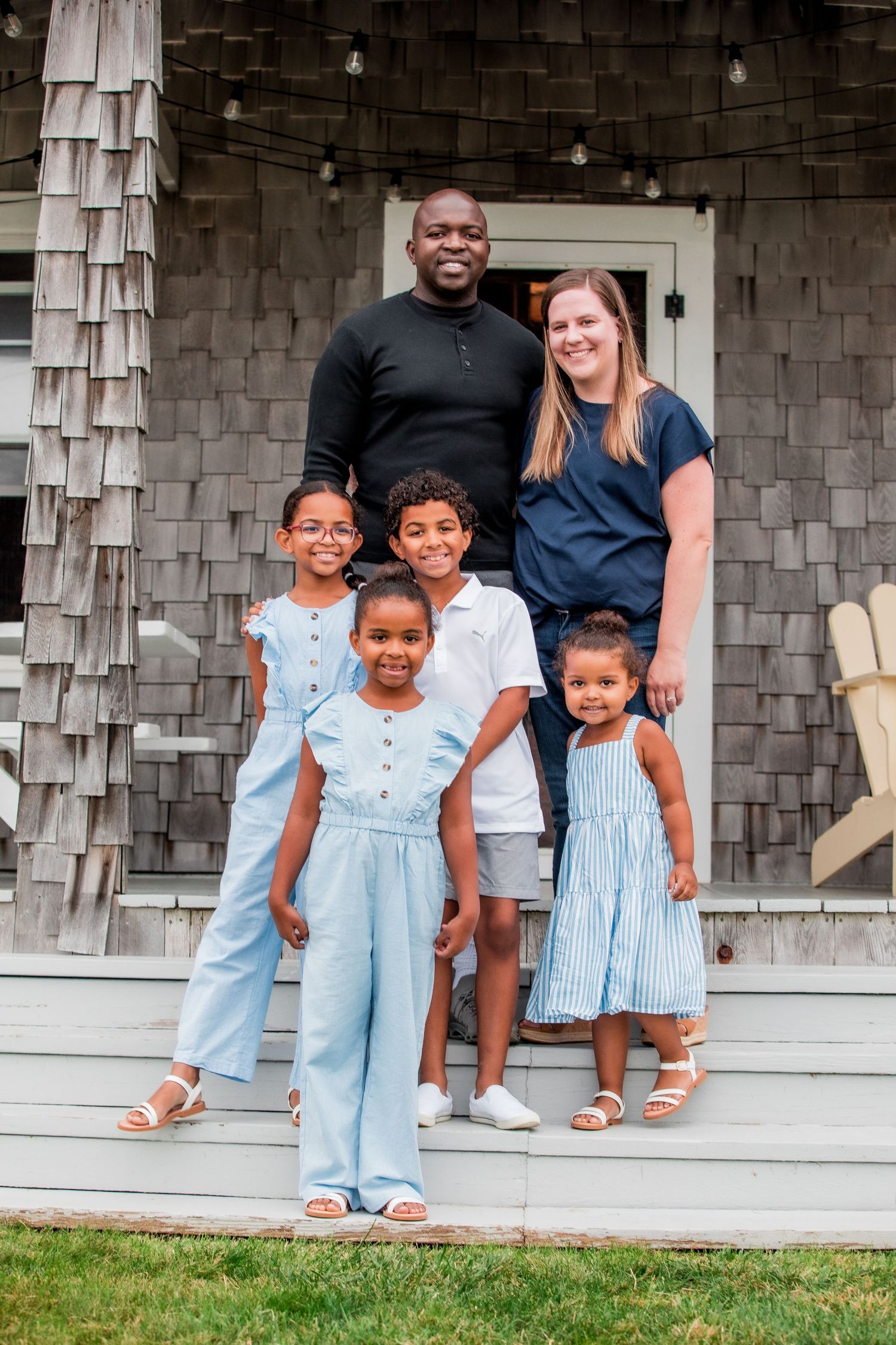 Multiracial family of six posing on porch steps of gray shingled home.