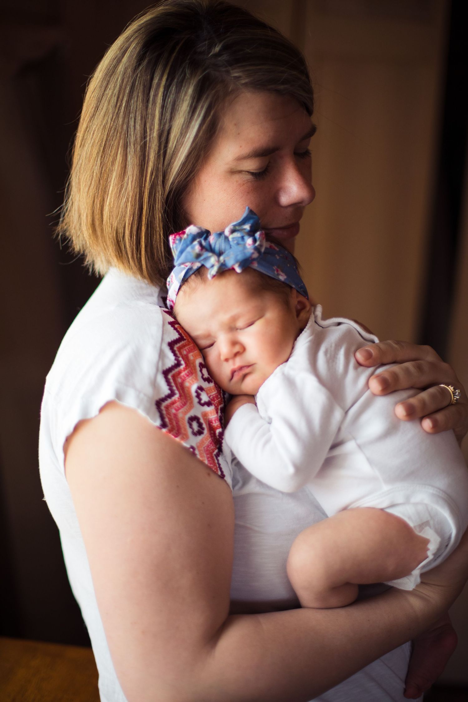 Mother with short hair tenderly holding sleeping newborn baby with blue bow headband.