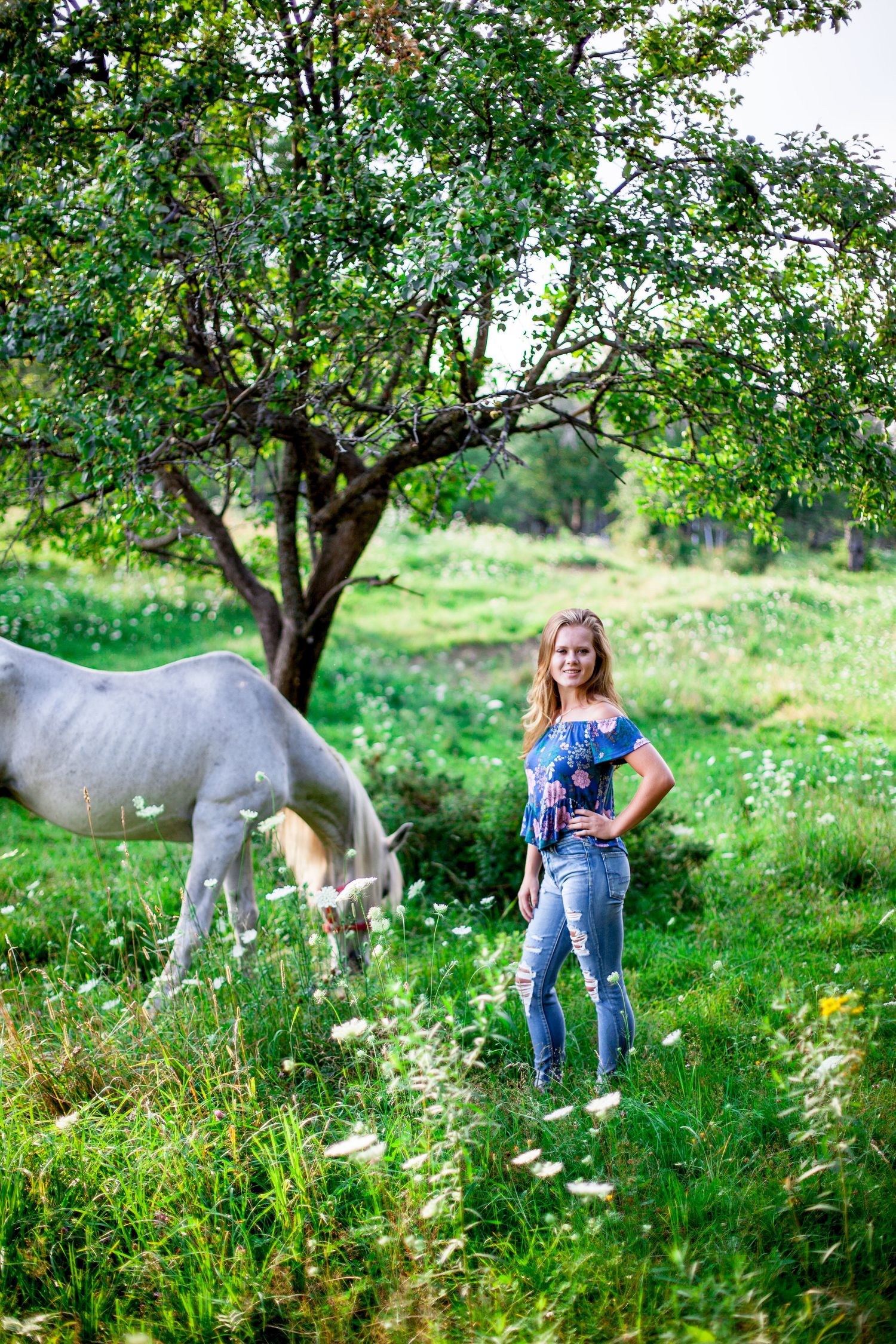 Young blonde woman standing in green meadow holding flowers with a white horse grazing under a tree nearby.