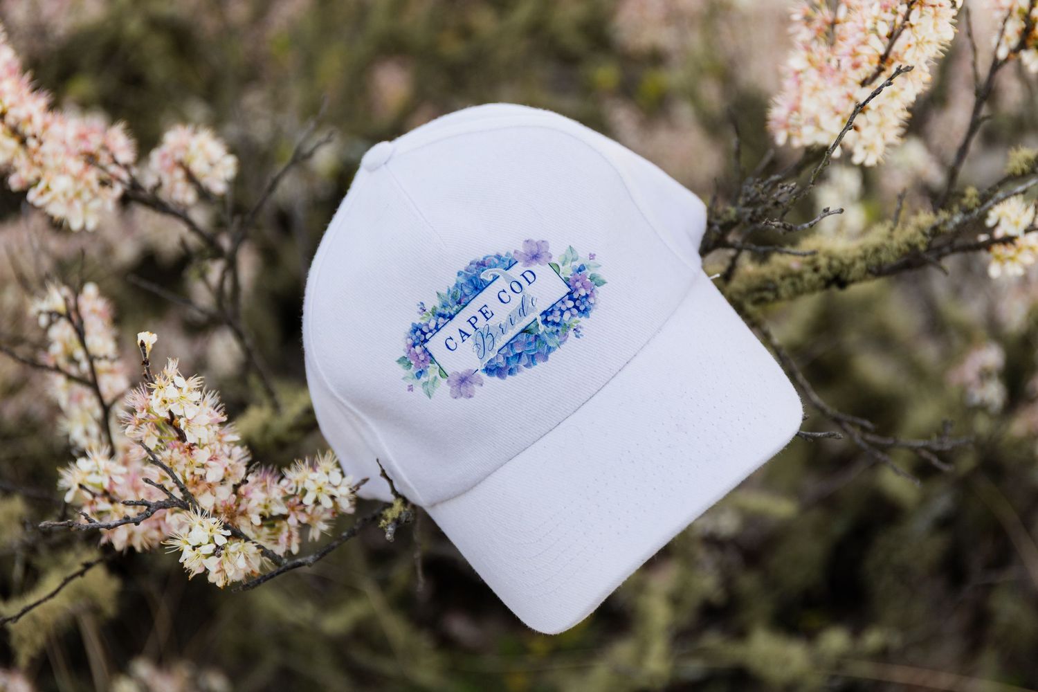 White baseball cap with floral Cape Cod embroidery resting on blooming spring branches.