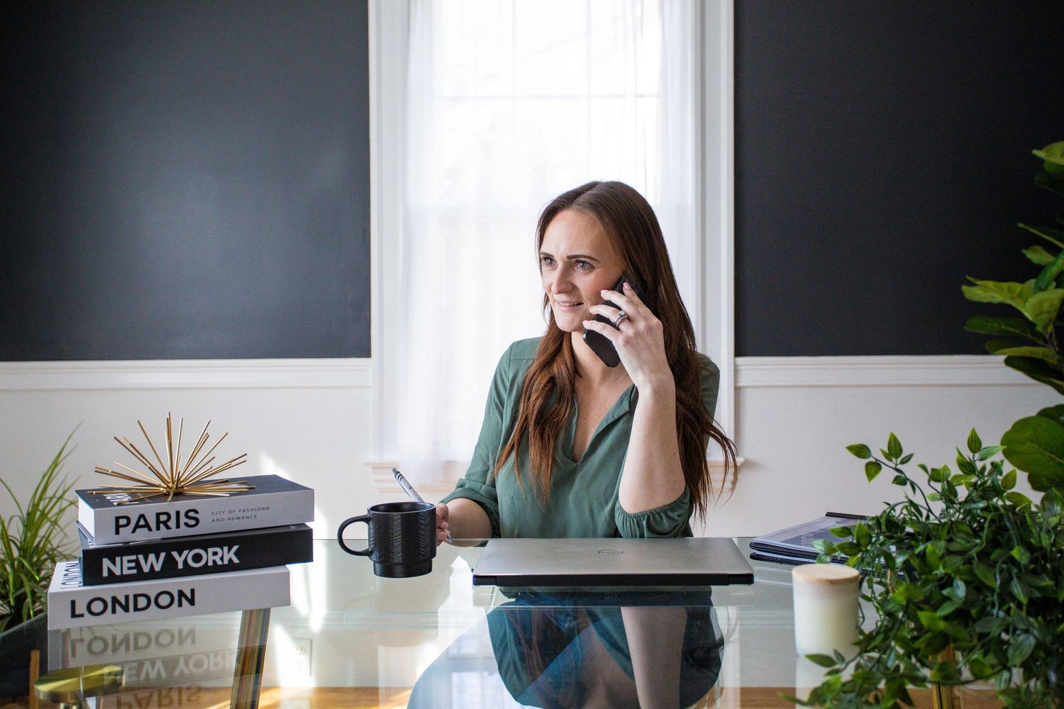Smiling woman talking on phone at glass desk with city-themed books and indoor plants.