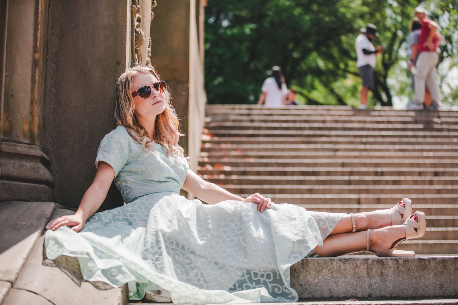Blonde woman in sunglasses and light blue midi dress relaxing on stone steps in Central Park.