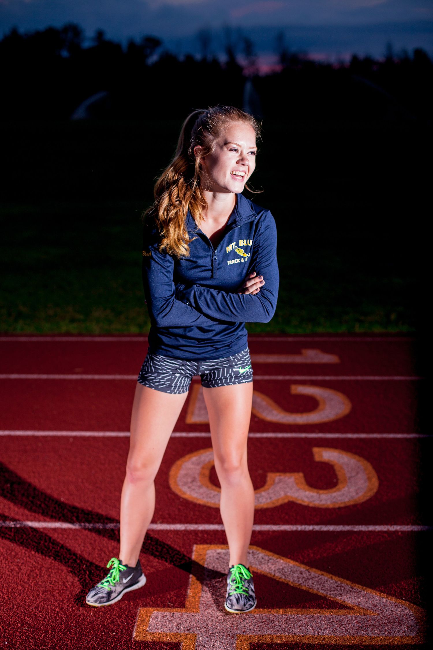 Female athlete standing confidently on a track at dusk wearing a blue team jacket.
