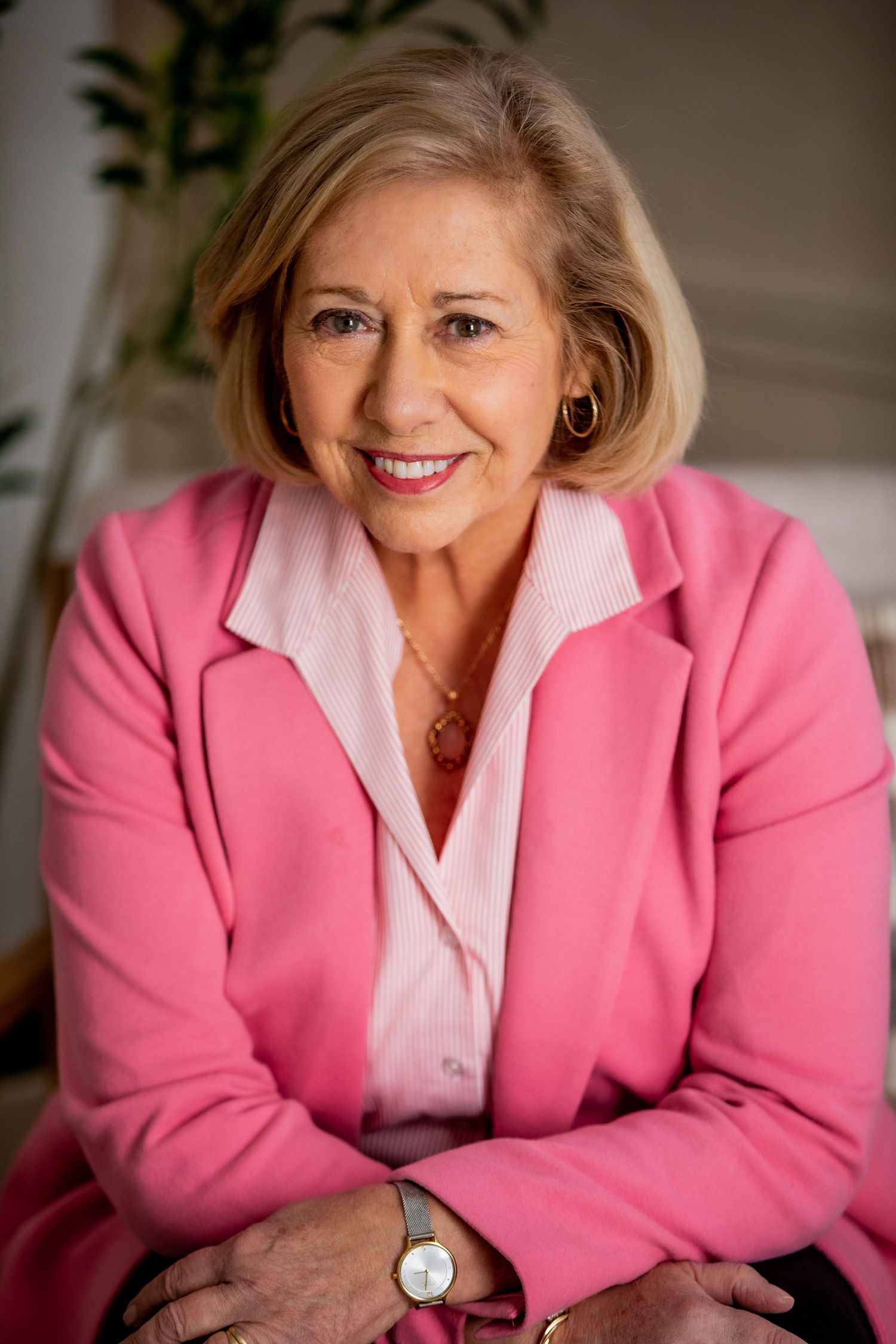 Smiling older woman in a pink blazer and blouse seated with arms crossed in a bright room.