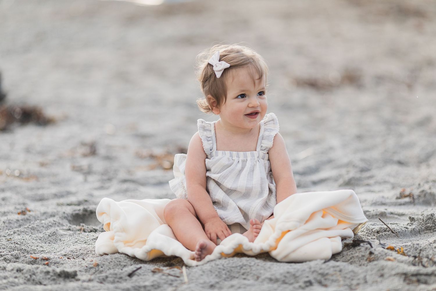 Adorable toddler girl in a gray romper sitting on sandy  Cape Cod beach with a blanket.