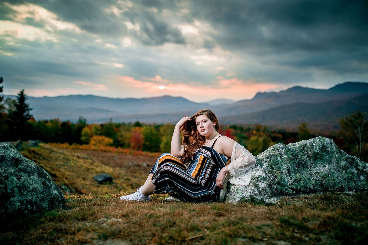 A young woman in a striped dress sits against a rock with a dramatic mountain sunset backdrop.