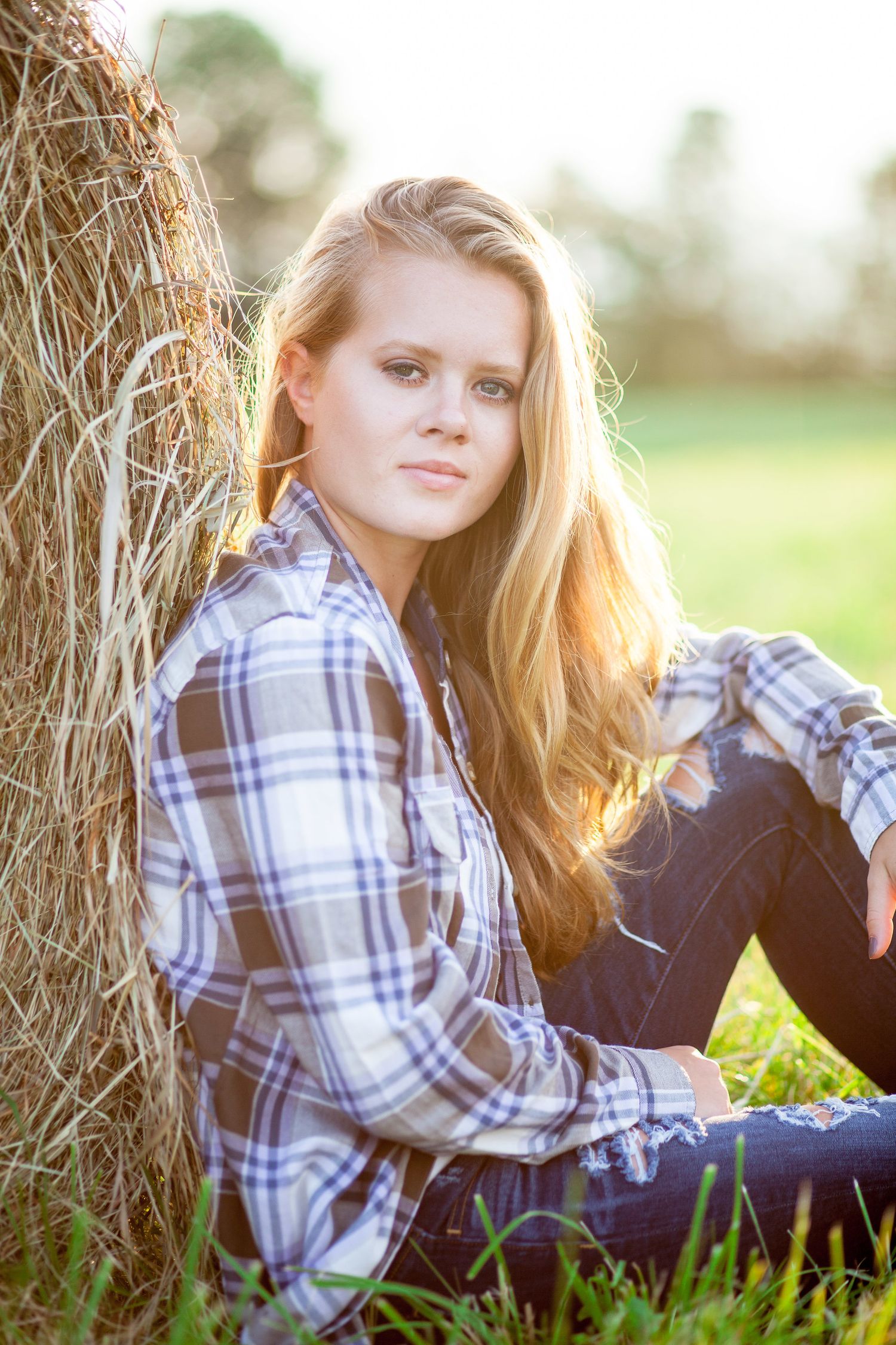 Blonde teenage girl in plaid shirt sitting against a hay bale in a sunny field.