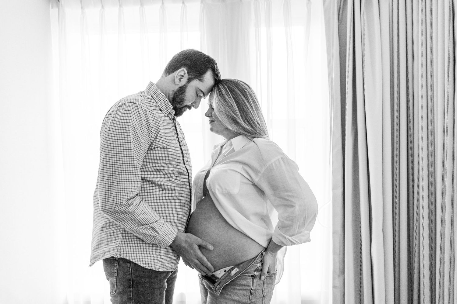 Black and white photo of expecting couple touching foreheads by bright window.