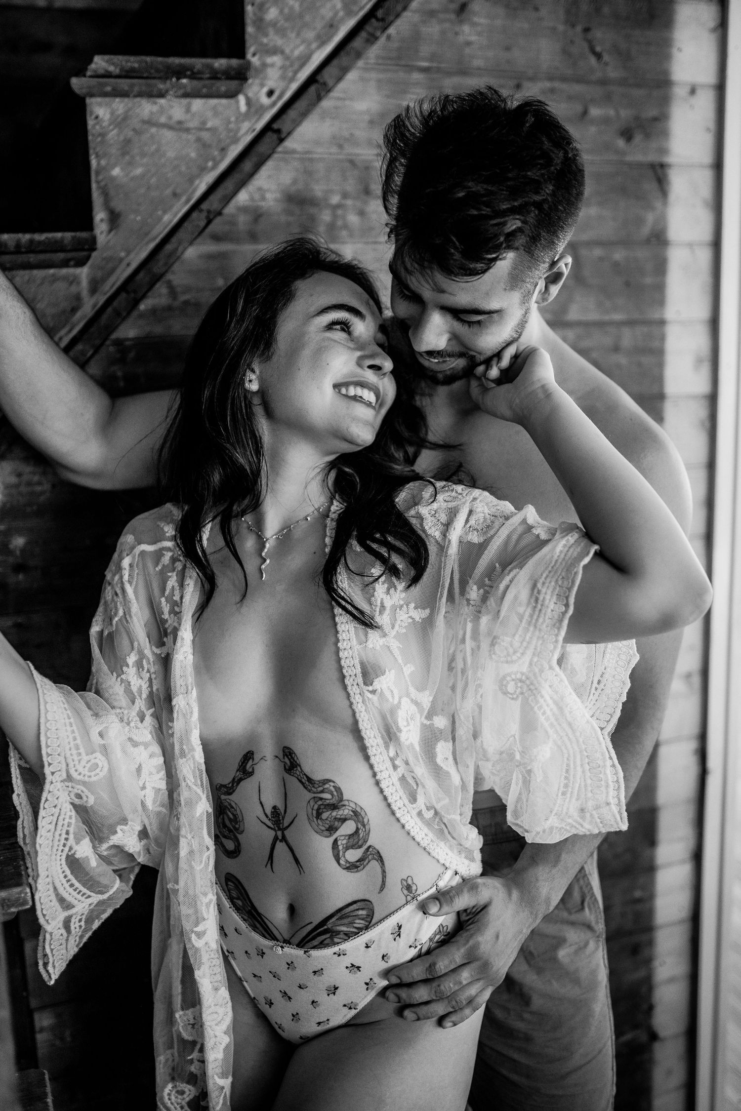 Black and white photo of a smiling couple embracing near wooden stairs, woman wearing lace robe with body tattoos.