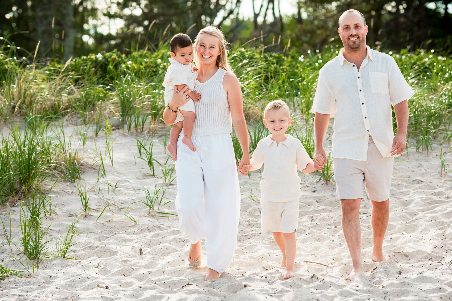 Happy family of four walking on a sandy beach, parents holding children's hands with green dunes in background.