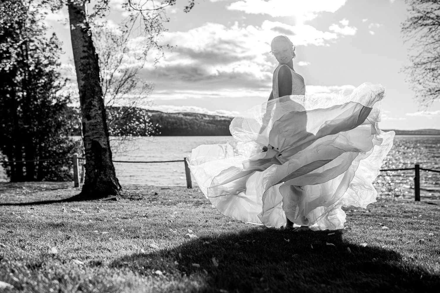 Black and white photo of bride twirling wedding dress by lakeside trees.