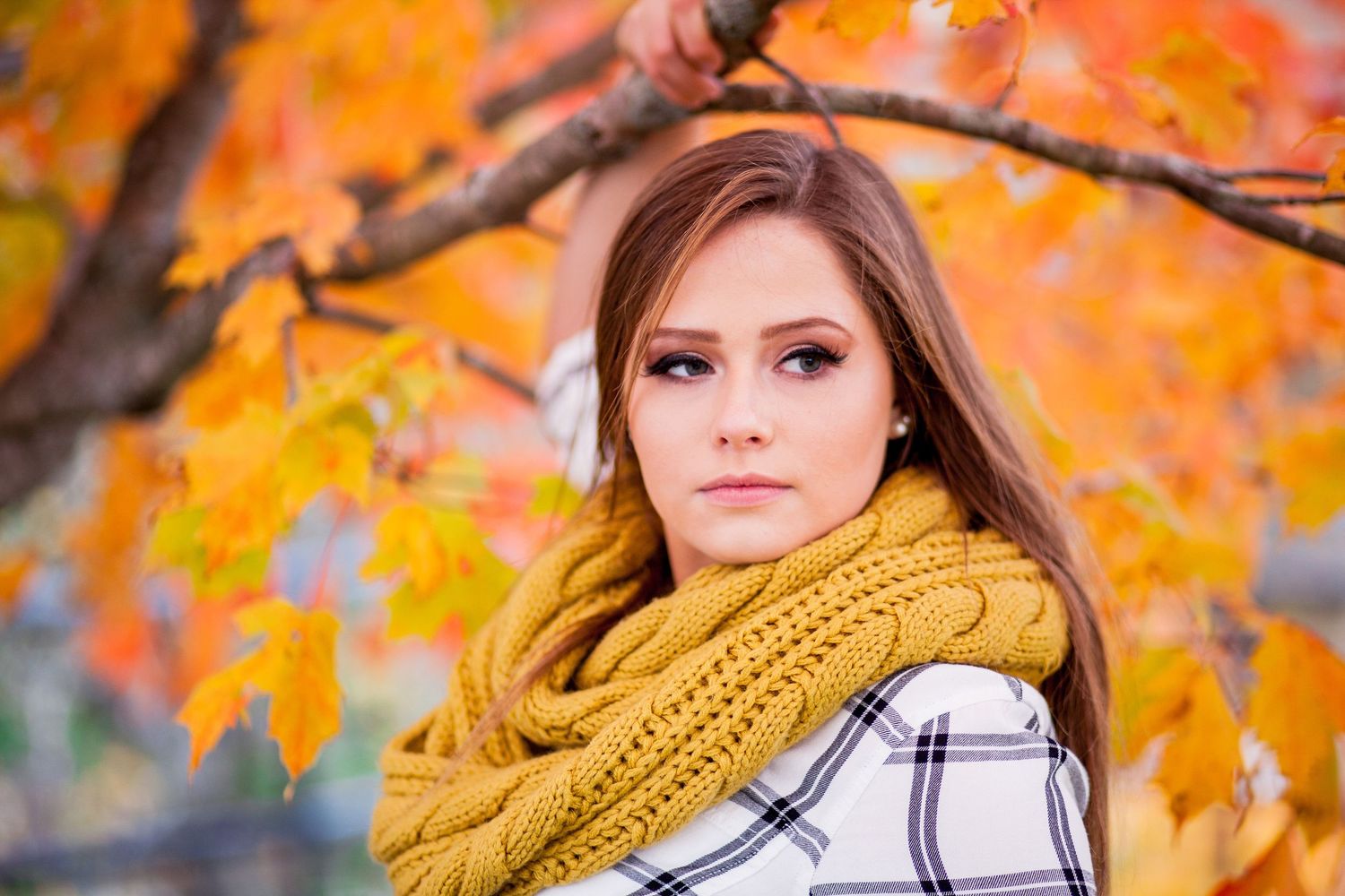 Close-up portrait of a young woman in yellow knit scarf and plaid coat surrounded by vibrant orange autumn leaves.